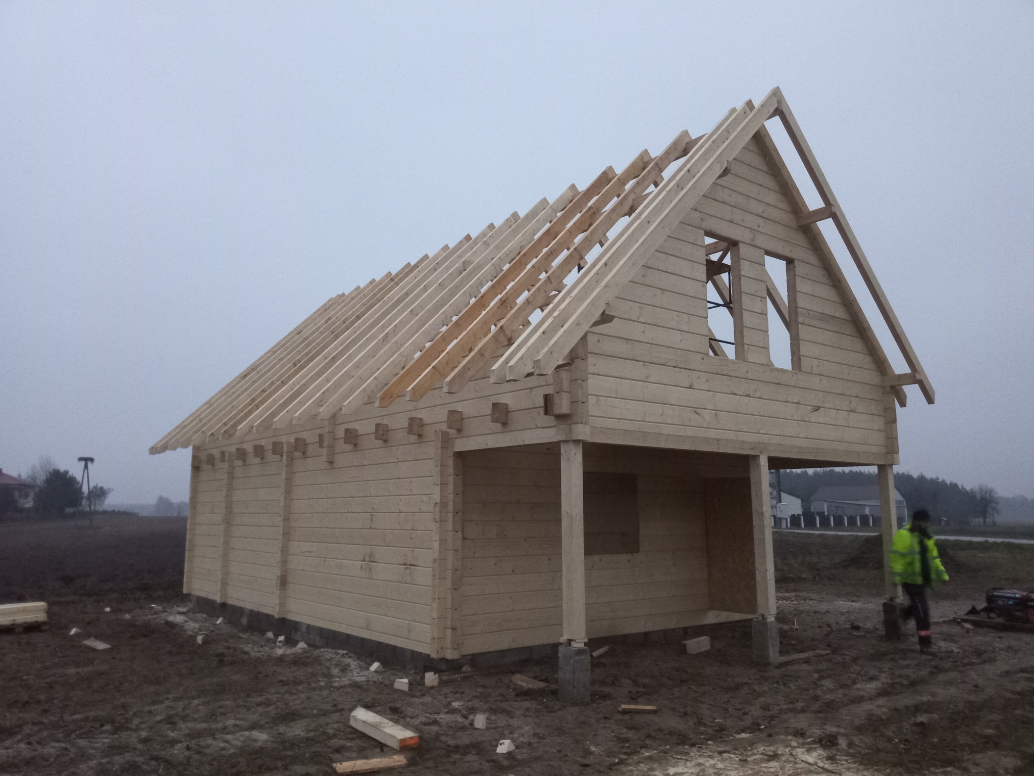 Surowy stan konstrukcji drewnianej budynku z widocznymi belkami stropowymi i szkieletem dachu na betonowych fundamentach, pracownik w odblaskowej kurtce w tle.
