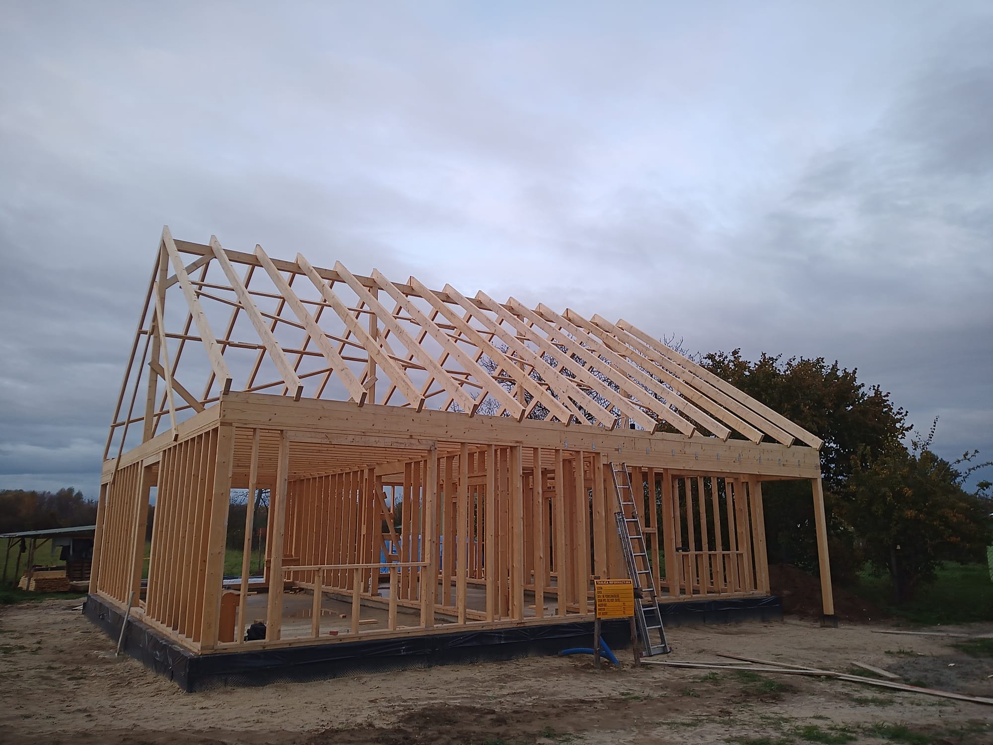 Konstrukcja domu w trakcie budowy, widoczne drewniane ramy ścian i dachu, drabina oparta o ścianę, tablica informacyjna budowy, pochmurne niebo w tle.