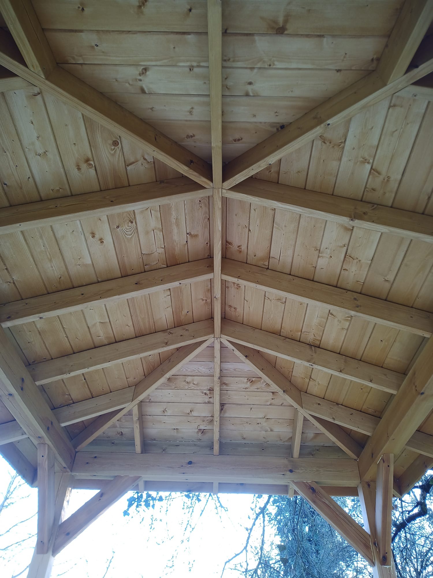 Wnętrze drewnianej altany ogrodowej z widoczną konstrukcją dachu, wykonaną z jasnego drewna, z deskami ułożonymi w jodełkę.