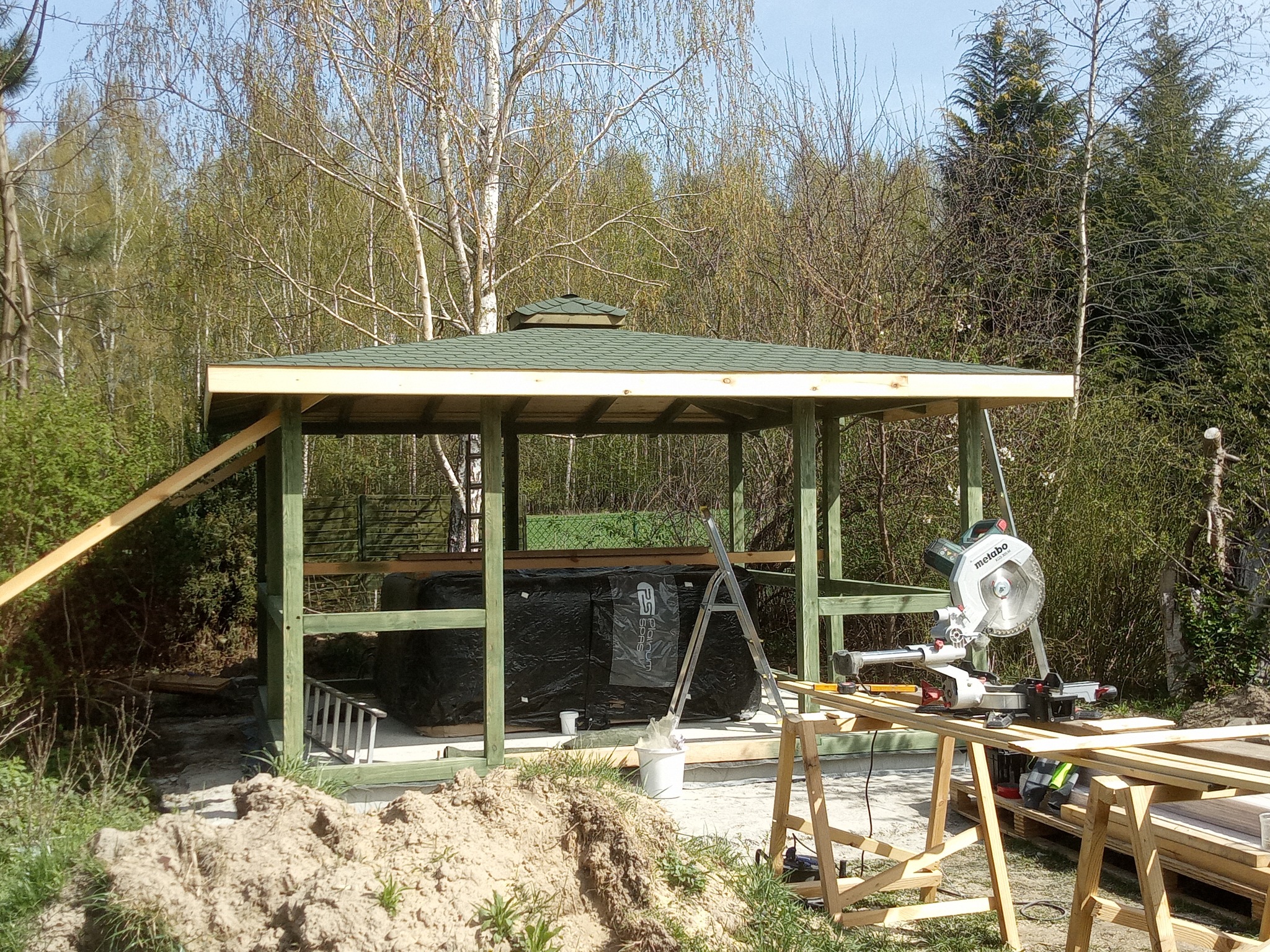 Konstrukcja altany w ogrodzie w trakcie budowy; widoczna zielona konstrukcja z dachem, piła Metabo na stole warsztatowym, w tle drzewa.