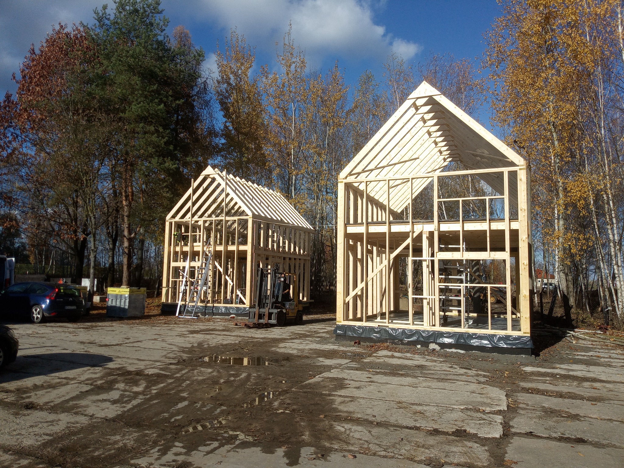 Dwa surowe szkielety domów z drewna na betonowej płycie, z widocznymi elementami konstrukcyjnymi i podnośnikiem w tle, otoczone drzewami i błękitnym niebem.