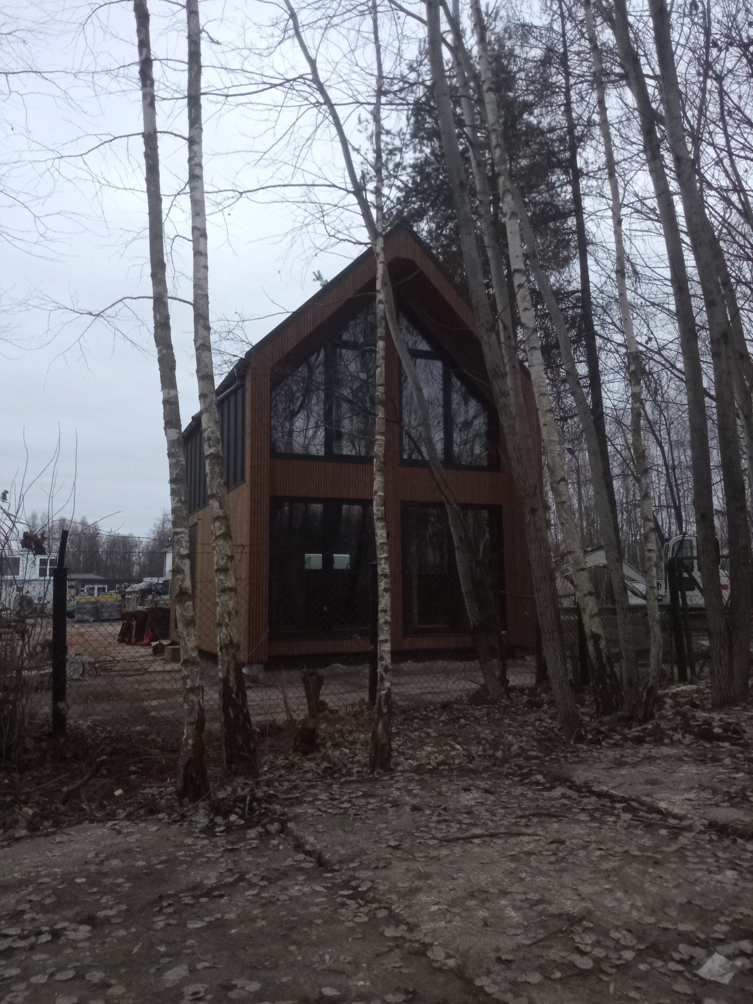 Nowoczesny dom z drewnianą elewacją i dużymi oknami, częściowo zasłonięty przez drzewa. Widoczne elementy placu budowy w tle.