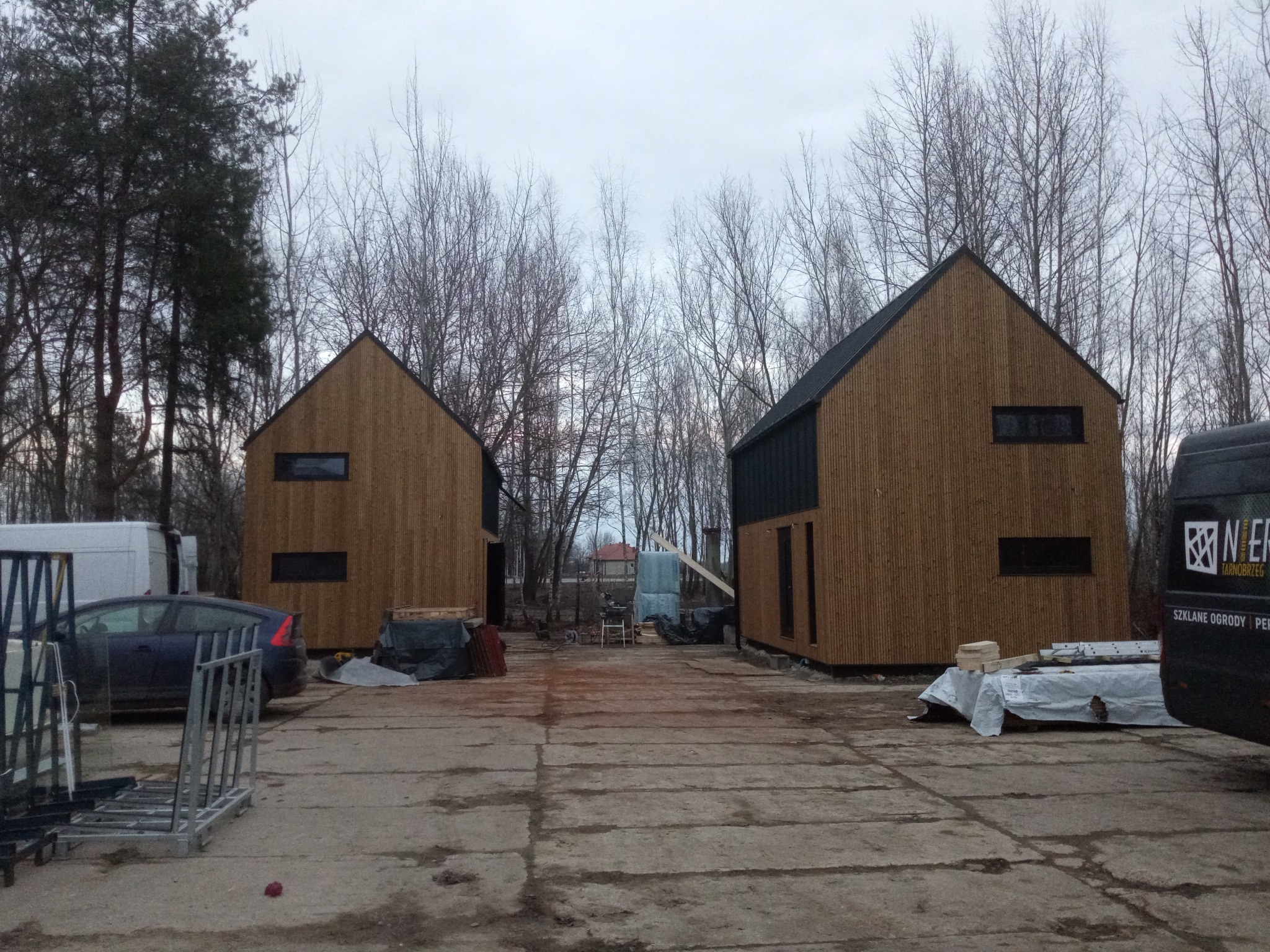 Dwa nowoczesne, drewniane domy z bali z czarnymi akcentami, ustawione symetrycznie na betonowym placu, otoczone drzewami bez liści i sprzętem budowlanym.