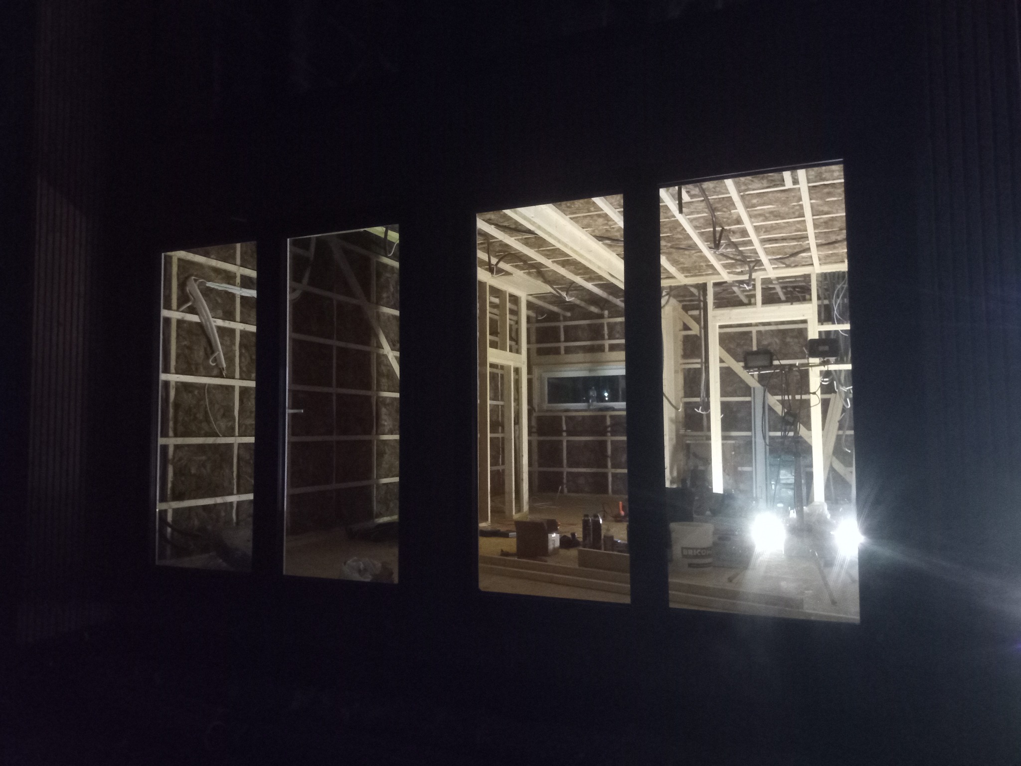 Wnętrze domu w trakcie budowy, widoczne przez okna. Konstrukcja szkieletowa z izolacją, narzędzia na podłodze, oświetlenie robocze. Ujęcie nocne z zewnątrz.