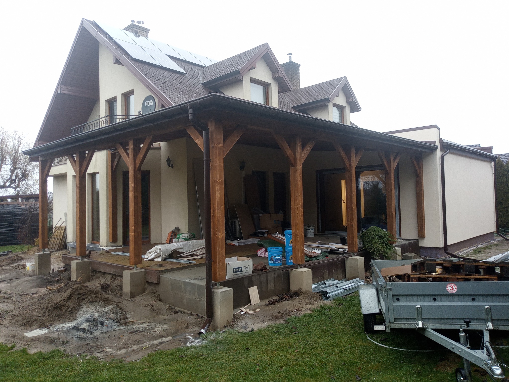 Dom z drewnianą konstrukcją werandy w trakcie budowy. Widoczne materiały budowlane, przyczepka i fragment ogrodu. Na dachu panele słoneczne.