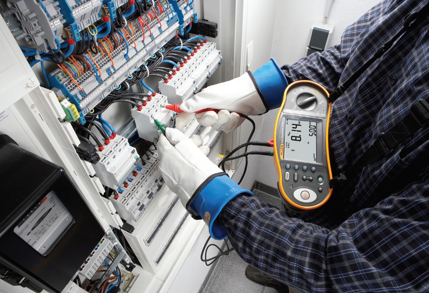 Elektryk w rękawicach ochronnych mierzy napięcie w rozdzielnicy elektrycznej za pomocą multimetru Fluke, widoczne przewody i zabezpieczenia