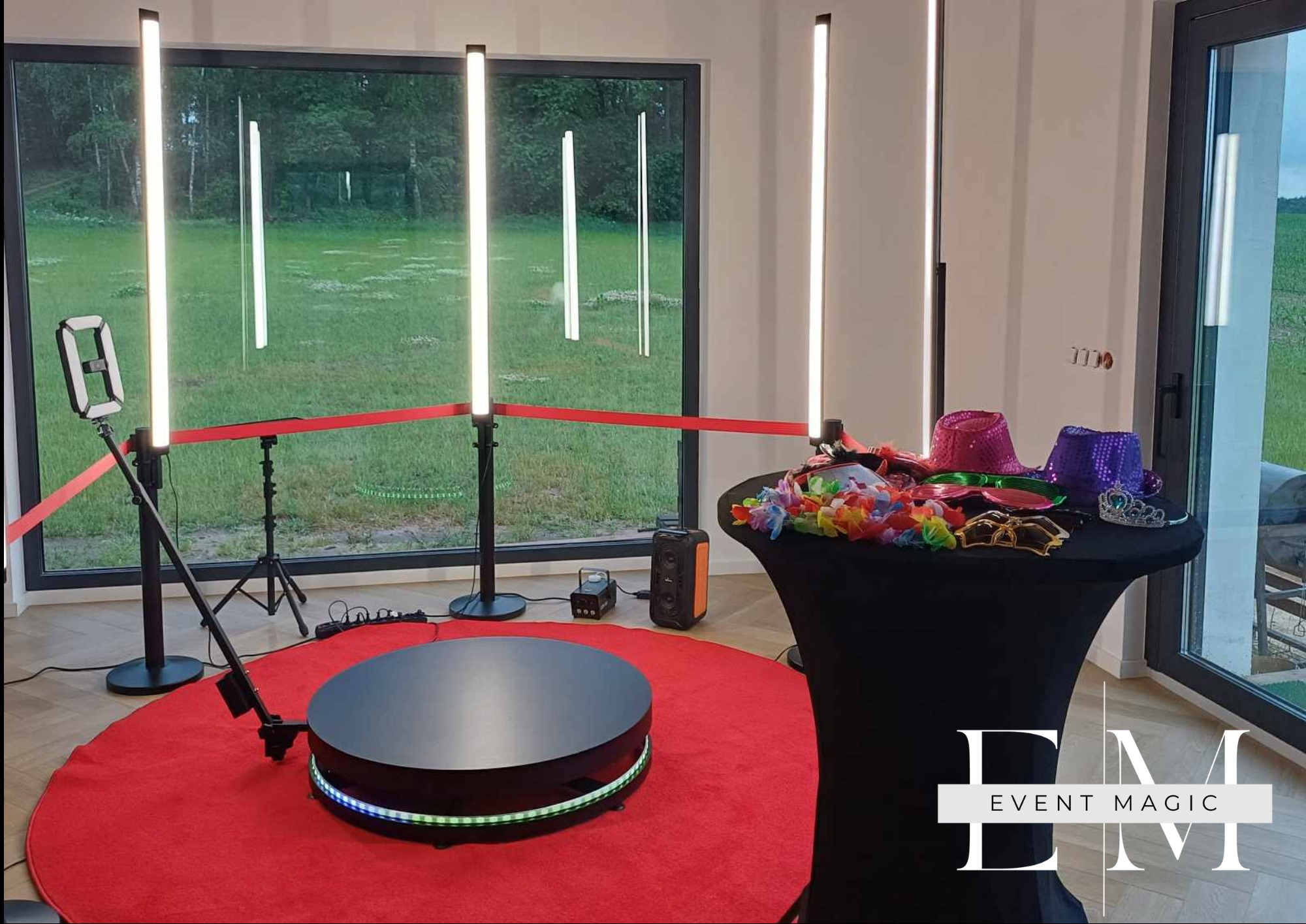 Nowoczesna fotobudka 360 na czerwonym dywanie, otoczona lampami LED i rekwizytami na stole. Widok na zewnątrz przez duże okno.