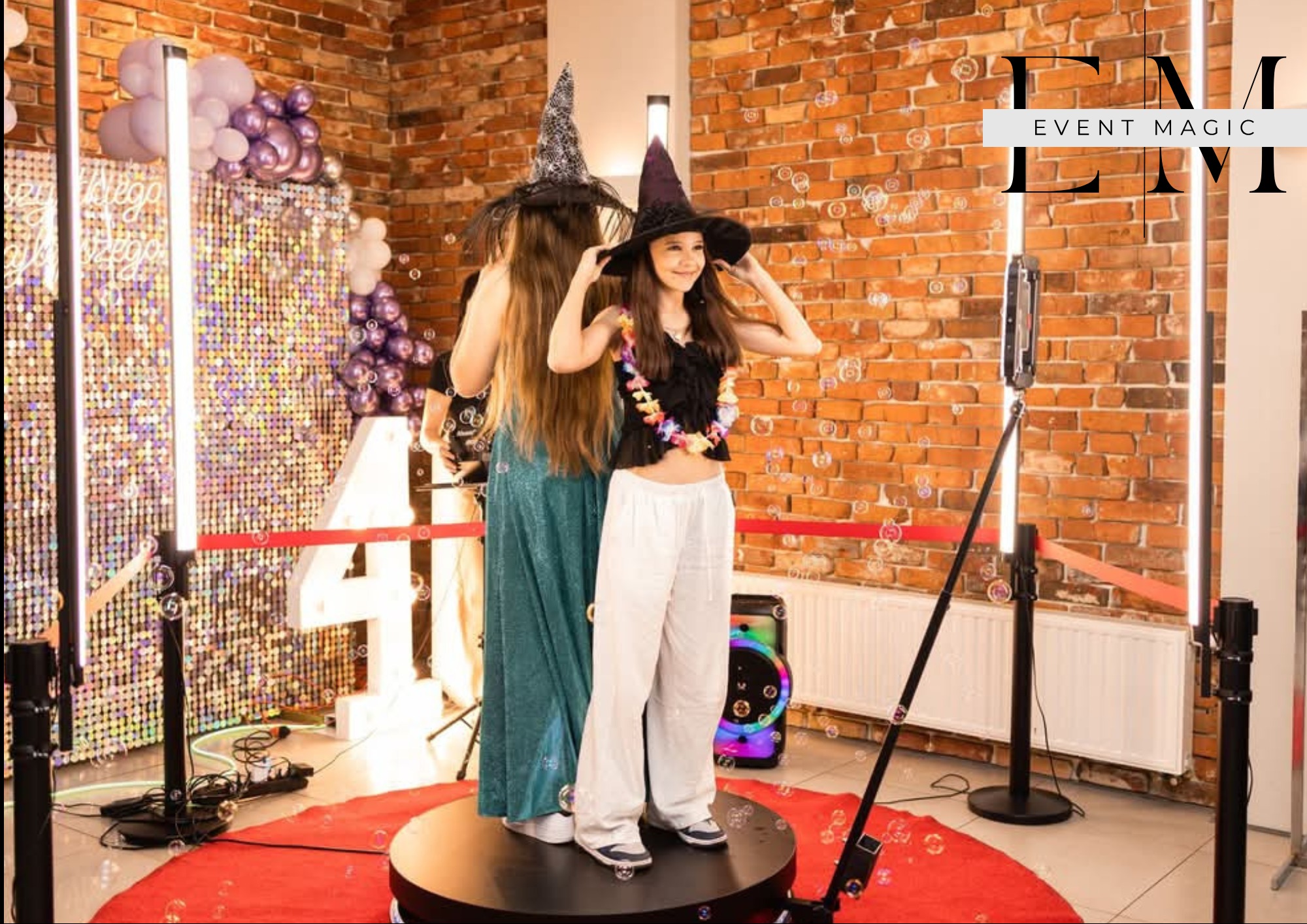 Dwie uśmiechnięte kobiety w czarowniczych kapeluszach pozują na obrotowej platformie z fotobudką w tle, dekoracje z balonów i ceglana ściana.