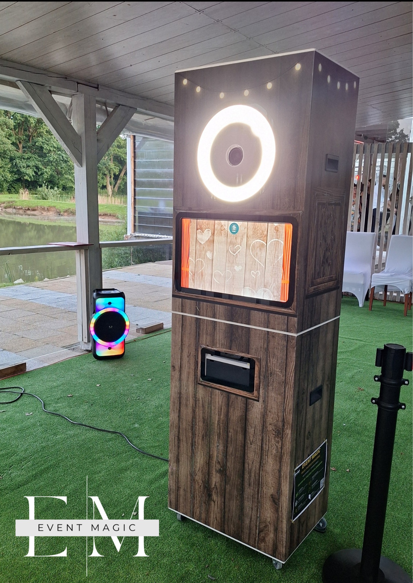 Mobilna fotobudka w drewnianej obudowie z oświetleniem LED, ustawiona na trawie obok kolorowego głośnika, w plenerze z widokiem na jezioro i altanę.