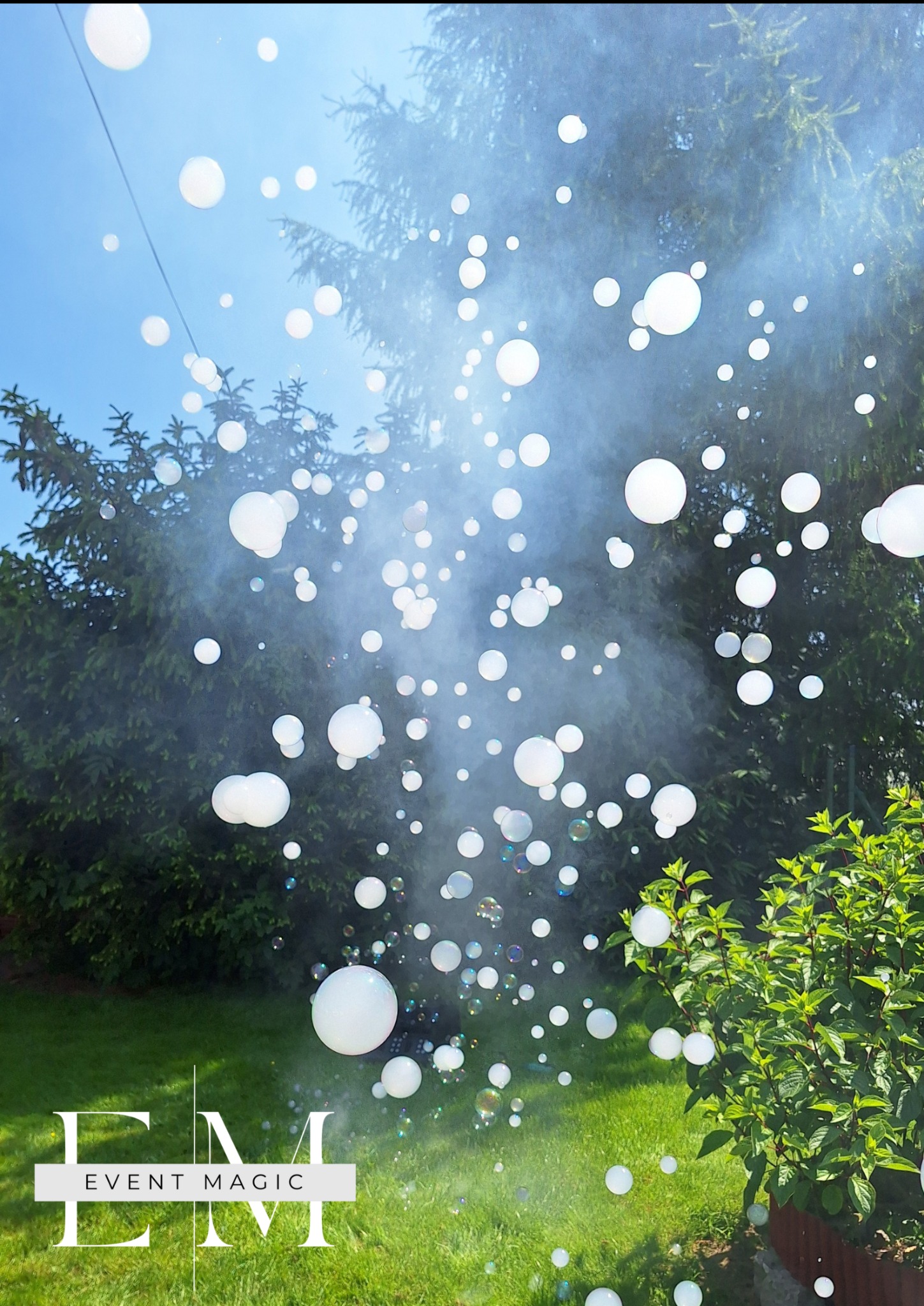 Efektowne bańki mydlane w powietrzu, generowane przez fotobudkę na tle zieleni i błękitnego nieba. Logo Event Magic w dolnej części kadru.