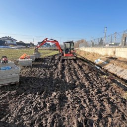 ZAKŁAD ROBÓT ZIEMNYCH Szymon Szolc - Czerwona minikoparka Kubota U36-4 pracuje na rozmokłym terenie, obok ułożone betonowe płyty na paletach i odkryty rów z płytami drenażowymi, w tle budynki.
