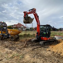ZAKŁAD ROBÓT ZIEMNYCH Szymon Szolc - Pomarańczowa koparka Kubota U50-5 z łyżką uniesioną nad ziemią, obok żółta wywrotka Terex na placu budowy z widocznymi domami w tle, pochmurne niebo.