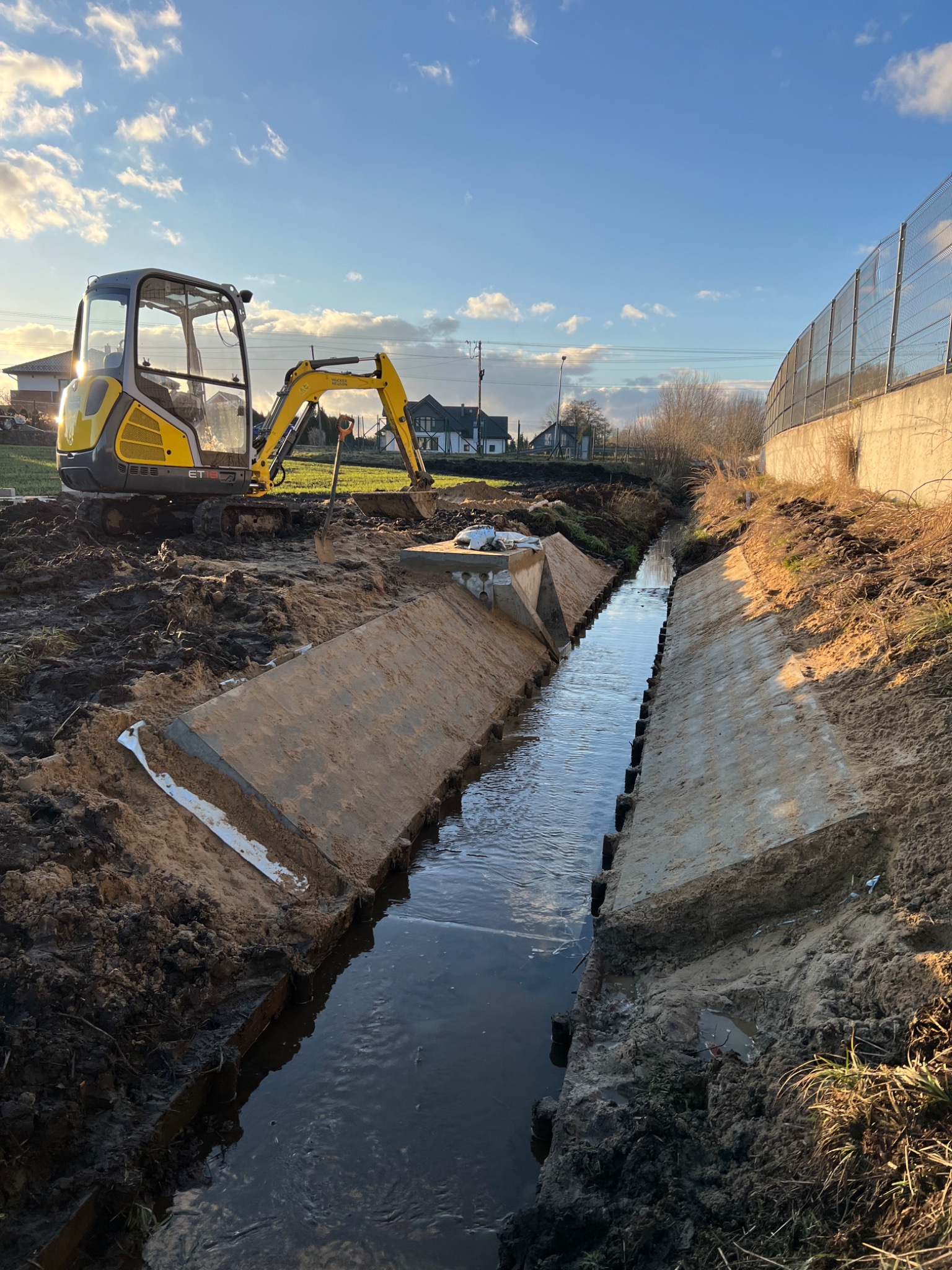 Żółta minikoparka stoi na grząskim terenie obok betonowego koryta wypełnionego wodą, w tle widoczne domy i błękitne niebo z chmurami.