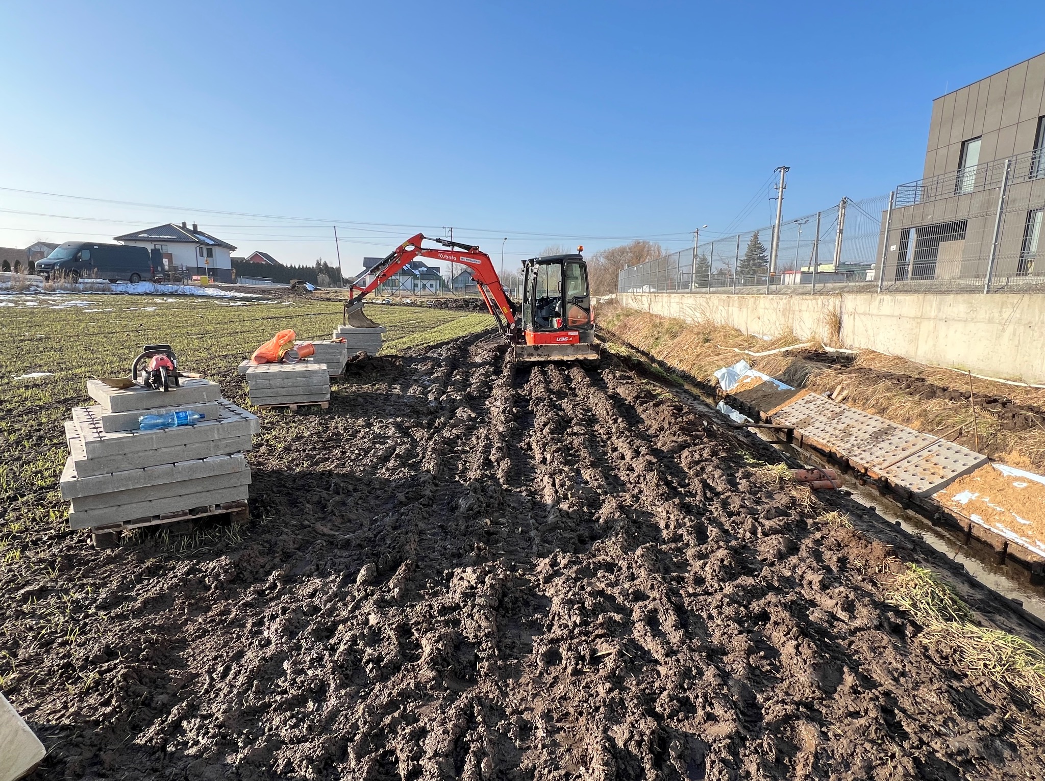 Czerwona minikoparka Kubota U36-4 pracuje na rozmokłym terenie, obok ułożone betonowe płyty na paletach i odkryty rów z płytami drenażowymi, w tle budynki.
