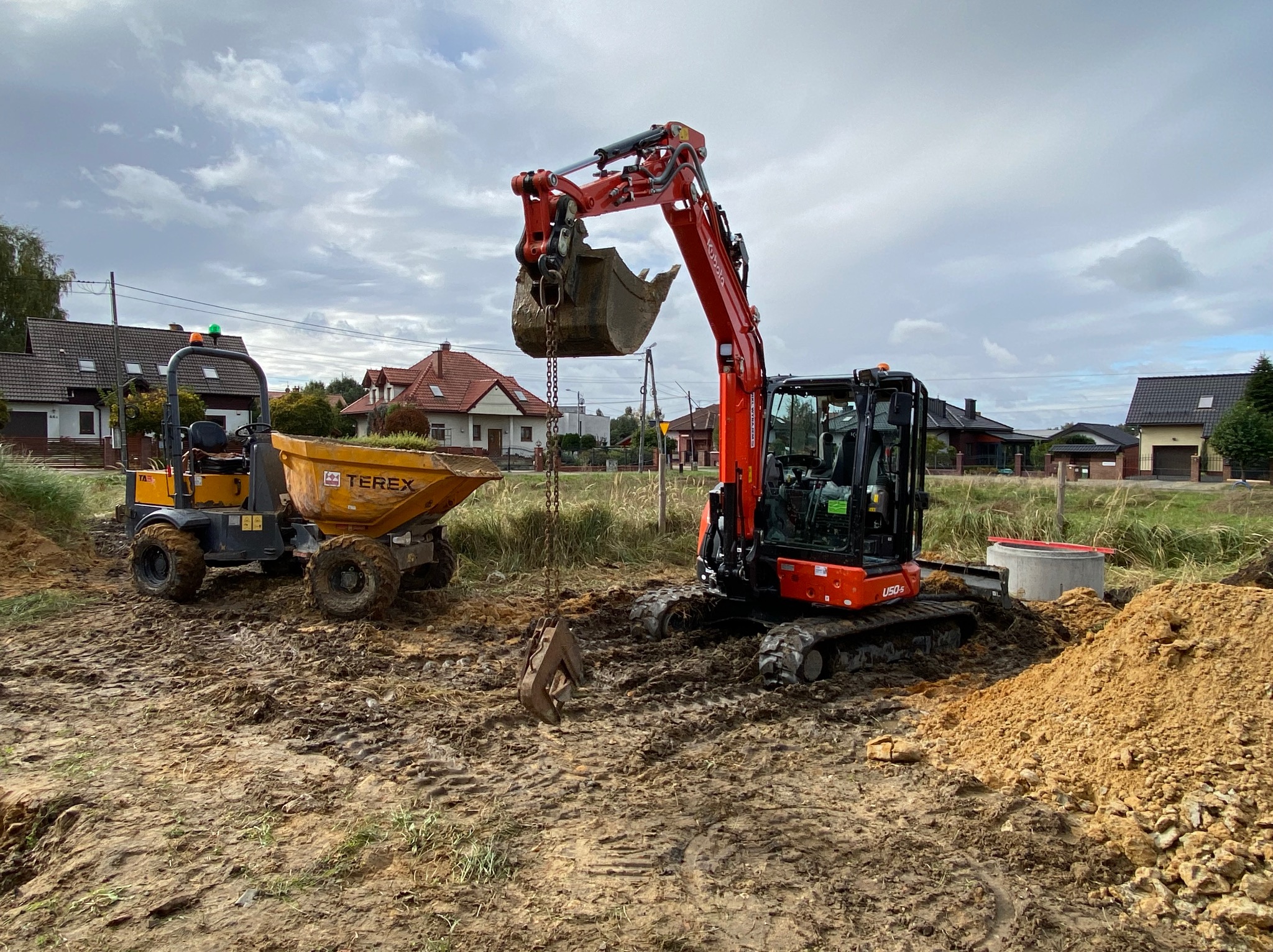 Pomarańczowa koparka Kubota U50-5 z łyżką uniesioną nad ziemią, obok żółta wywrotka Terex na placu budowy z widocznymi domami w tle, pochmurne niebo.