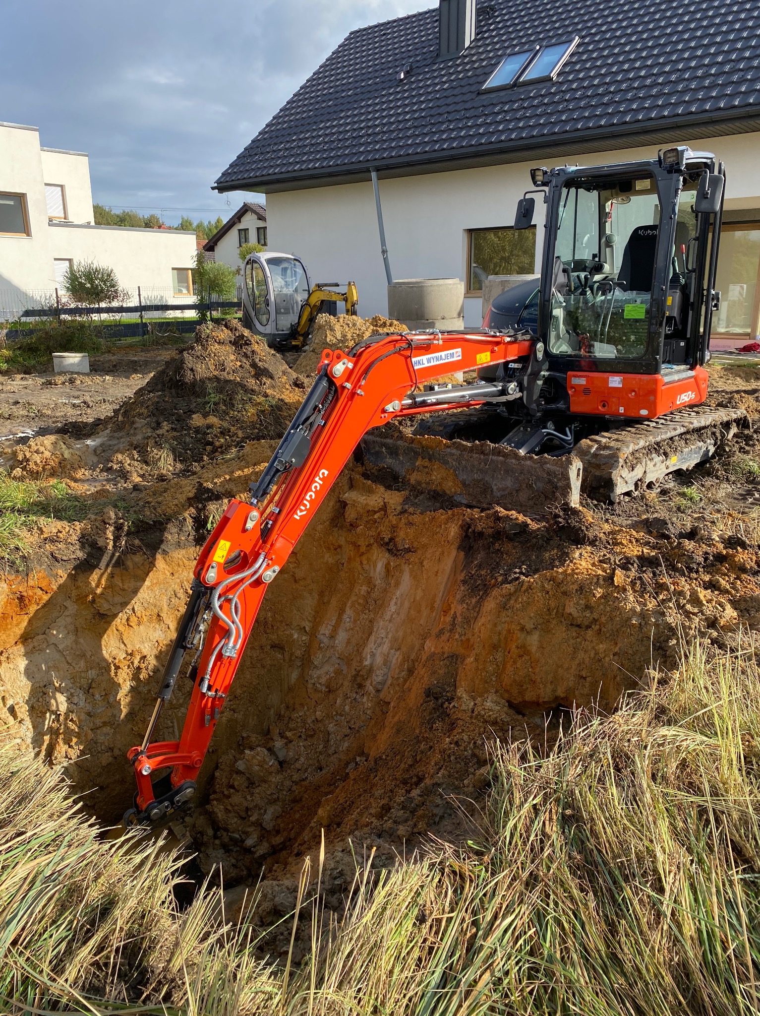 Pomarańczowa mini koparka Kubota U50-5 w trakcie prac ziemnych, widoczny głęboki wykop w gliniastej ziemi, w tle dom jednorodzinny i druga, mniejsza koparka.