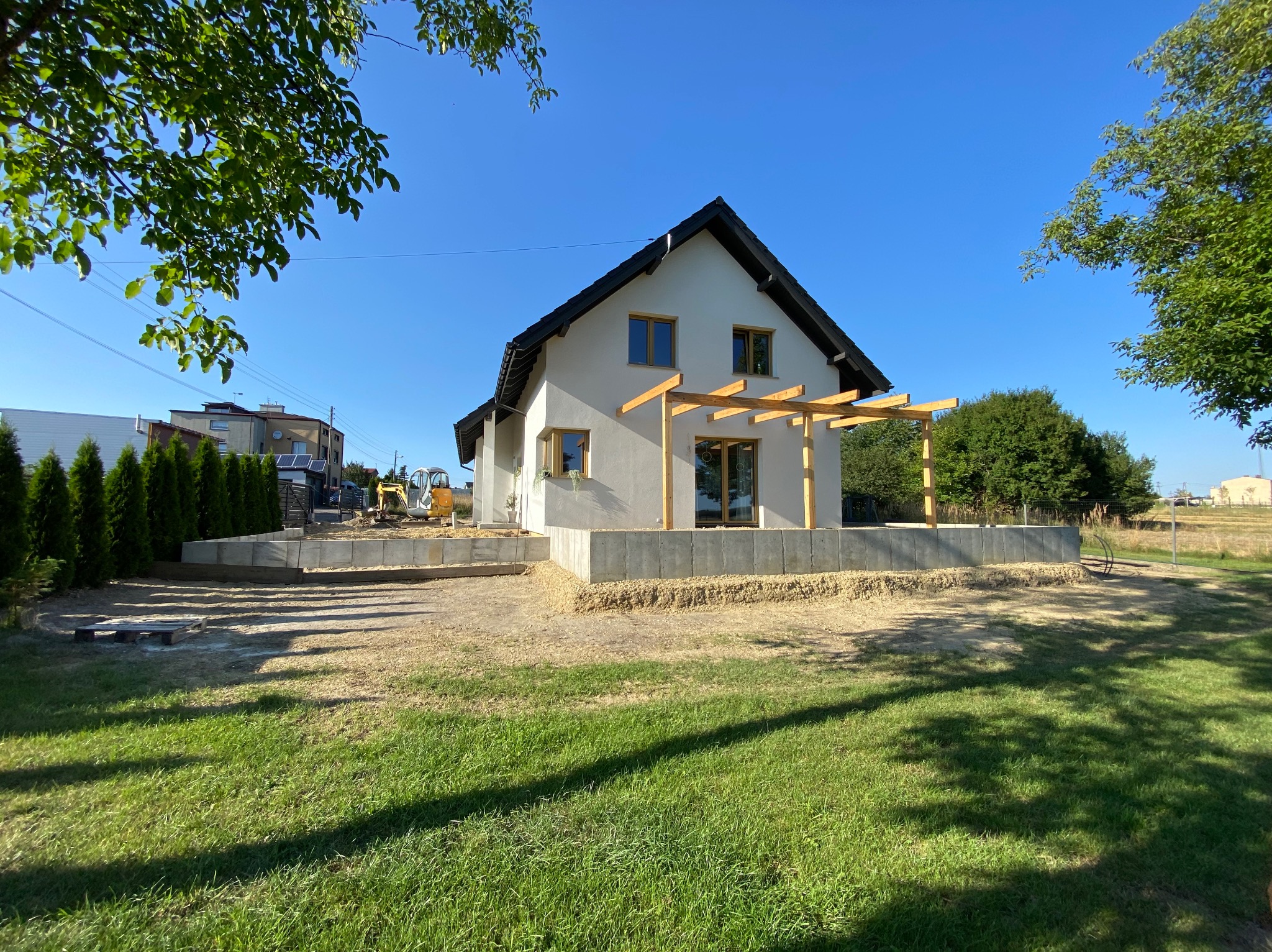 Nowoczesny dom jednorodzinny z jasną elewacją i drewnianą pergolą w trakcie budowy, widoczny fundament z bloczków betonowych i żółta koparka w tle.