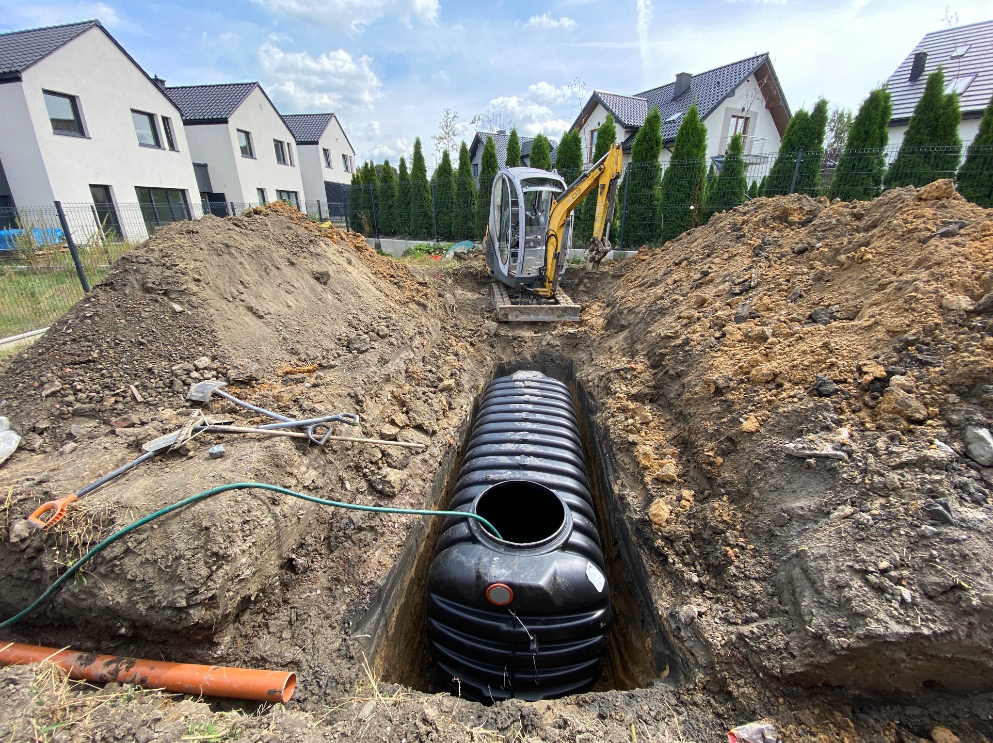 Czarny zbiornik z tworzywa sztucznego umieszczony w wykopie, widoczna mała koparka na tle nowoczesnych domów jednorodzinnych.