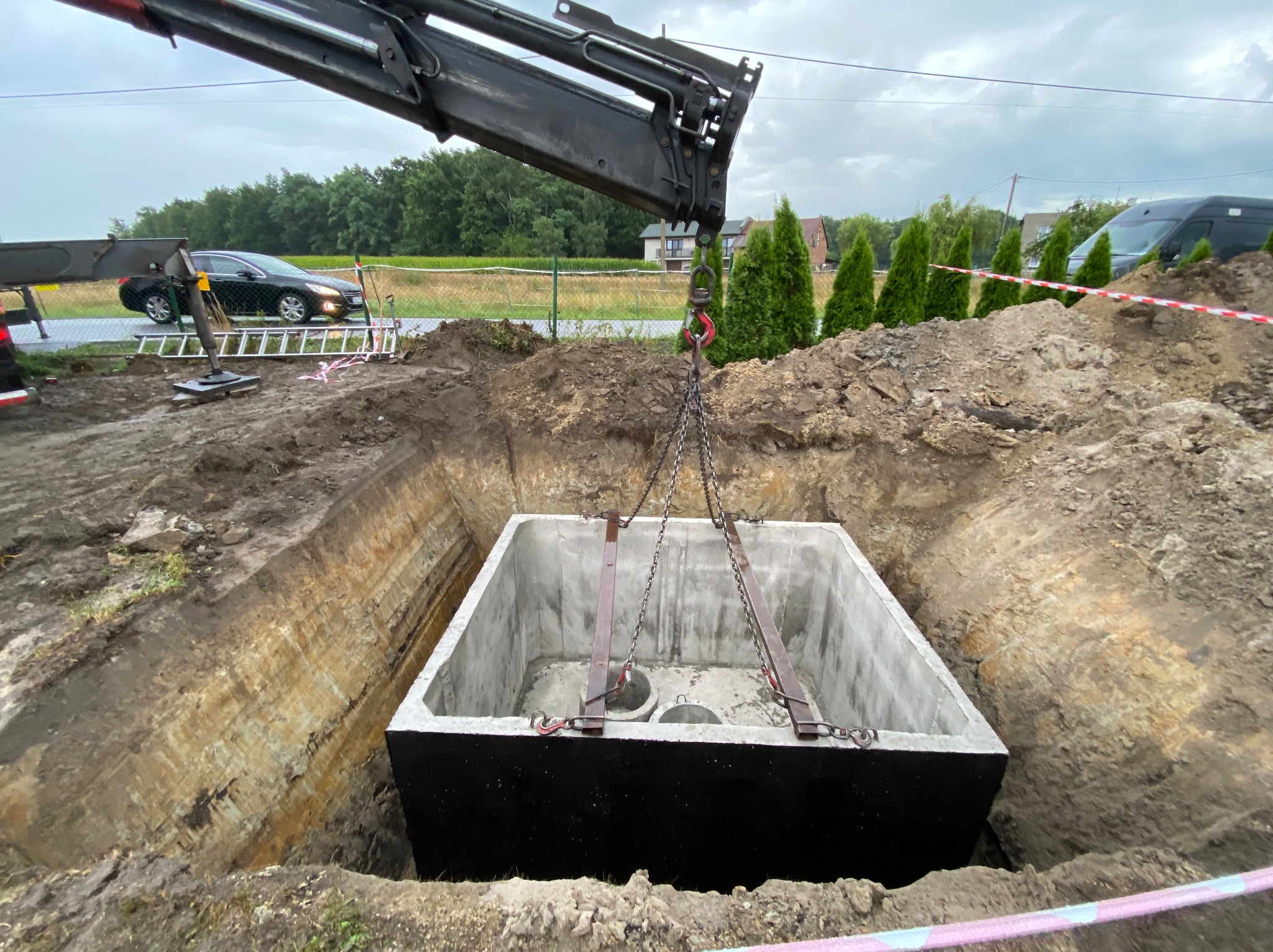 Betonowy zbiornik, prawdopodobnie szambo, opuszczany do wykopu za pomocą dźwigu; widoczne zabezpieczenia wykopu i otoczenie placu budowy.