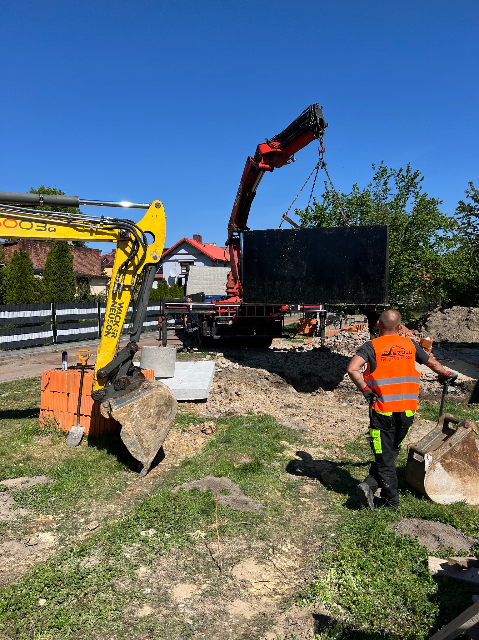 Montaż betonowego szamba za pomocą dźwigu na budowie. W tle widoczna żółta mini koparka i pracownik w kamizelce odblaskowej.