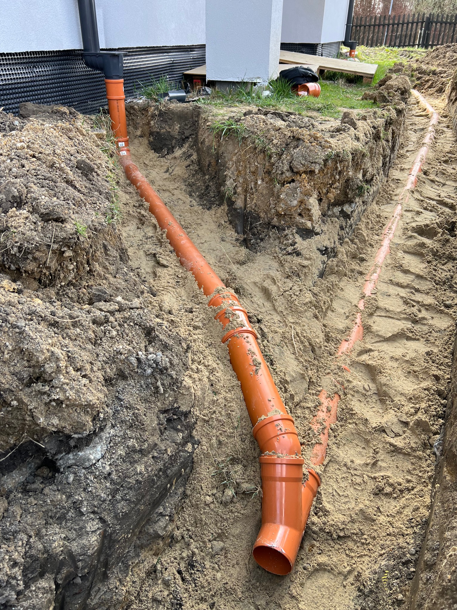 Pomarańczowe rury kanalizacyjne ułożone w wykopie ziemnym, widoczne połączenia i odgałęzienia. Prace instalacyjne w gruncie.