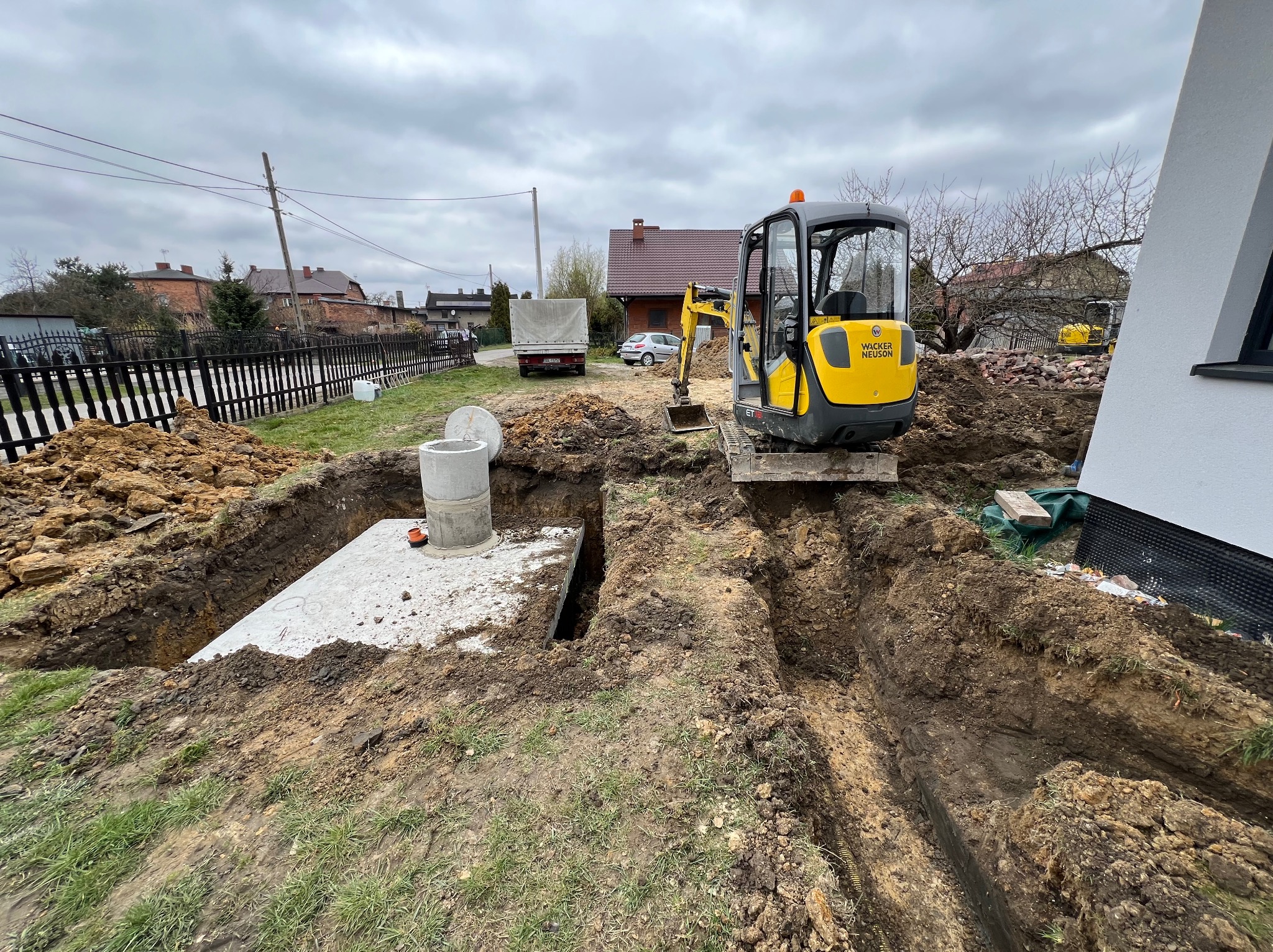 Żółta koparka Wacker Neuson przy wykopie pod szambo betonowe z elementami betonowymi na dnie, w tle domy i pochmurne niebo.