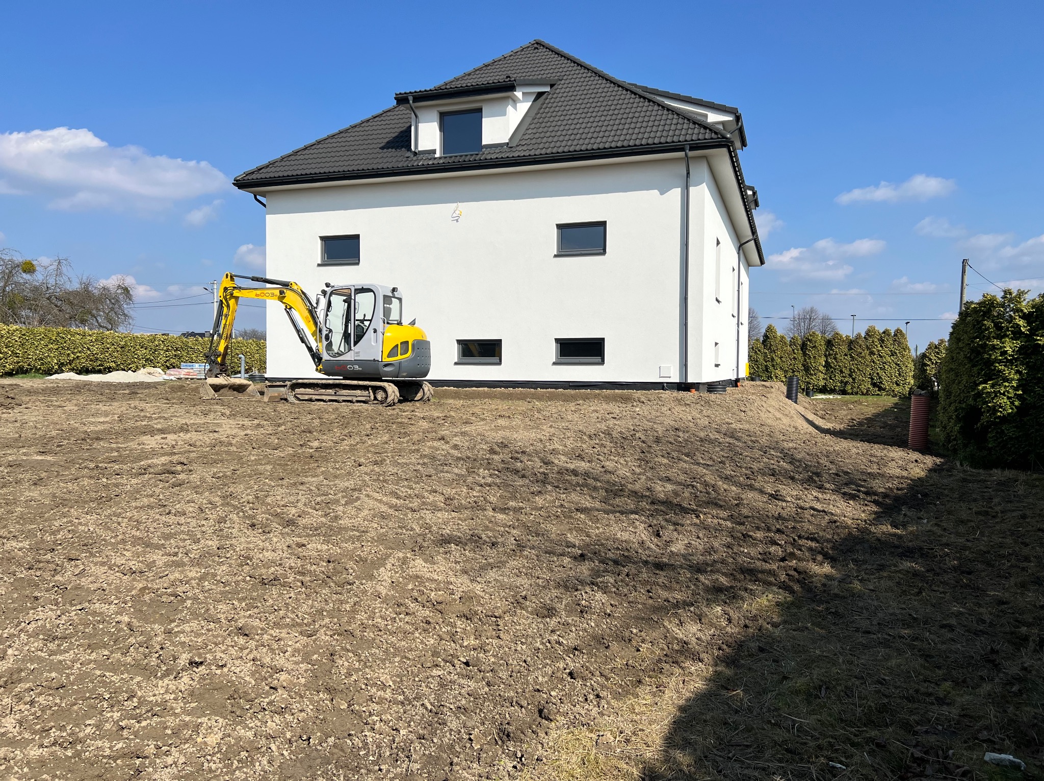 Żółta mini koparka na przygotowanym terenie budowy, obok nowoczesnego, białego domu z ciemną dachówką w słoneczny dzień. Widoczne rury drenażowe.