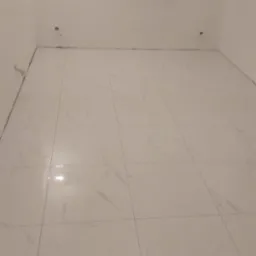 Wykończony pokój z białymi, marmurkowymi płytkami podłogowymi, z widocznymi przyłączami do armatury na ścianie.