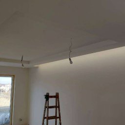 OdNowa FIRMA OGÓLNOBUDOWLANA BEATA KOWALCZYK - Wnętrze pokoju z pomalowanymi na biało ścianami i sufitem, z widocznymi elementami oświetlenia w suficie podwieszanym i wiszącymi żarówkami na kablach, drewniana drabina oparta o ścianę.