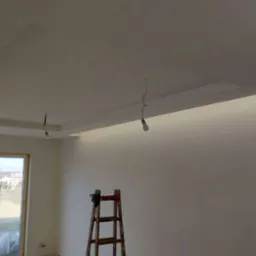 Wnętrze pokoju z pomalowanymi na biało ścianami i sufitem, z widocznymi elementami oświetlenia w suficie podwieszanym i wiszącymi żarówkami na kablach, drewniana drabina oparta o ścianę.