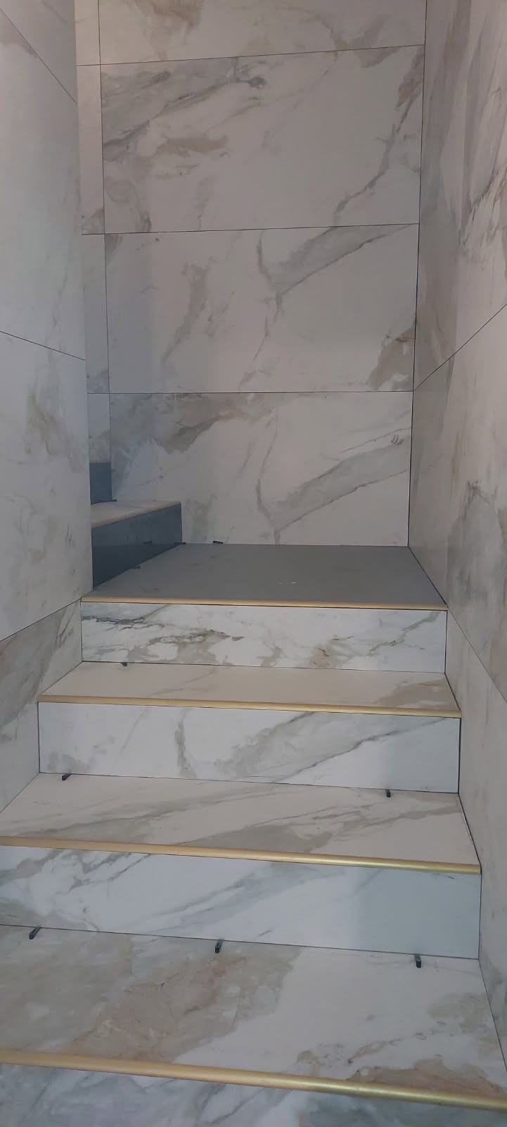 Schody wyłożone płytkami imitującymi marmur z zamontowanymi profilami schodowymi w kolorze złotym, widoczne klipsy poziomujące płytki.