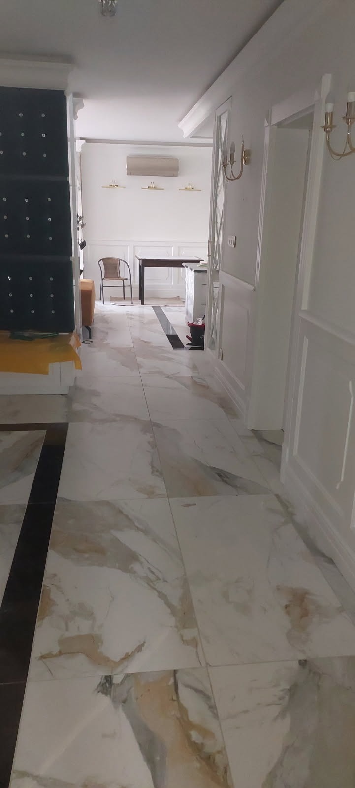 Wykończone wnętrze z marmurową podłogą w odcieniach bieli i beżu, białe ściany z dekoracyjnymi listwami, widoczne meble w tle, w tym czarna tapicerowana ściana z kryształkami i stół.