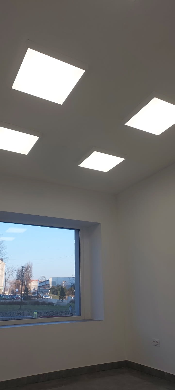 Wnętrze pomieszczenia po remoncie, widoczne cztery kwadratowe panele LED w suficie, okno z widokiem na zewnątrz i szare płytki podłogowe.
