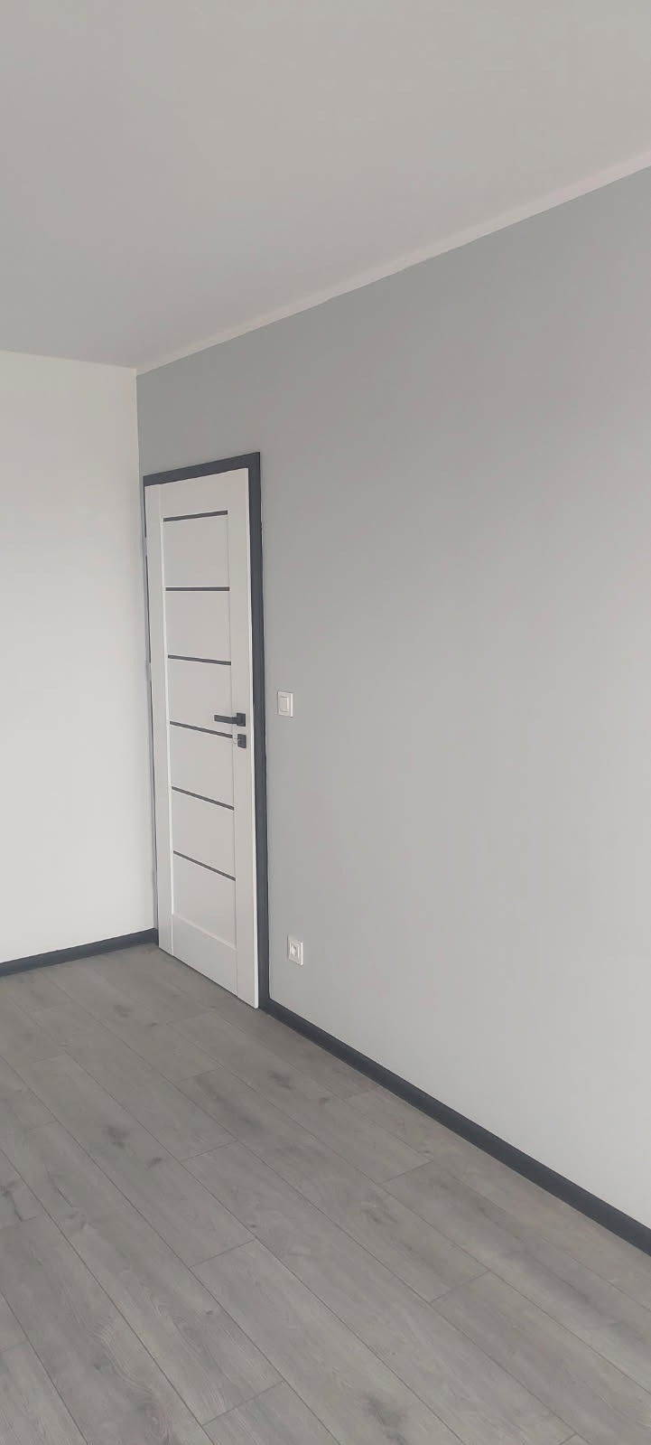 Pokój po remoncie: szare ściany, panele podłogowe w odcieniach szarości, białe drzwi z szarymi akcentami i szare listwy przypodłogowe.