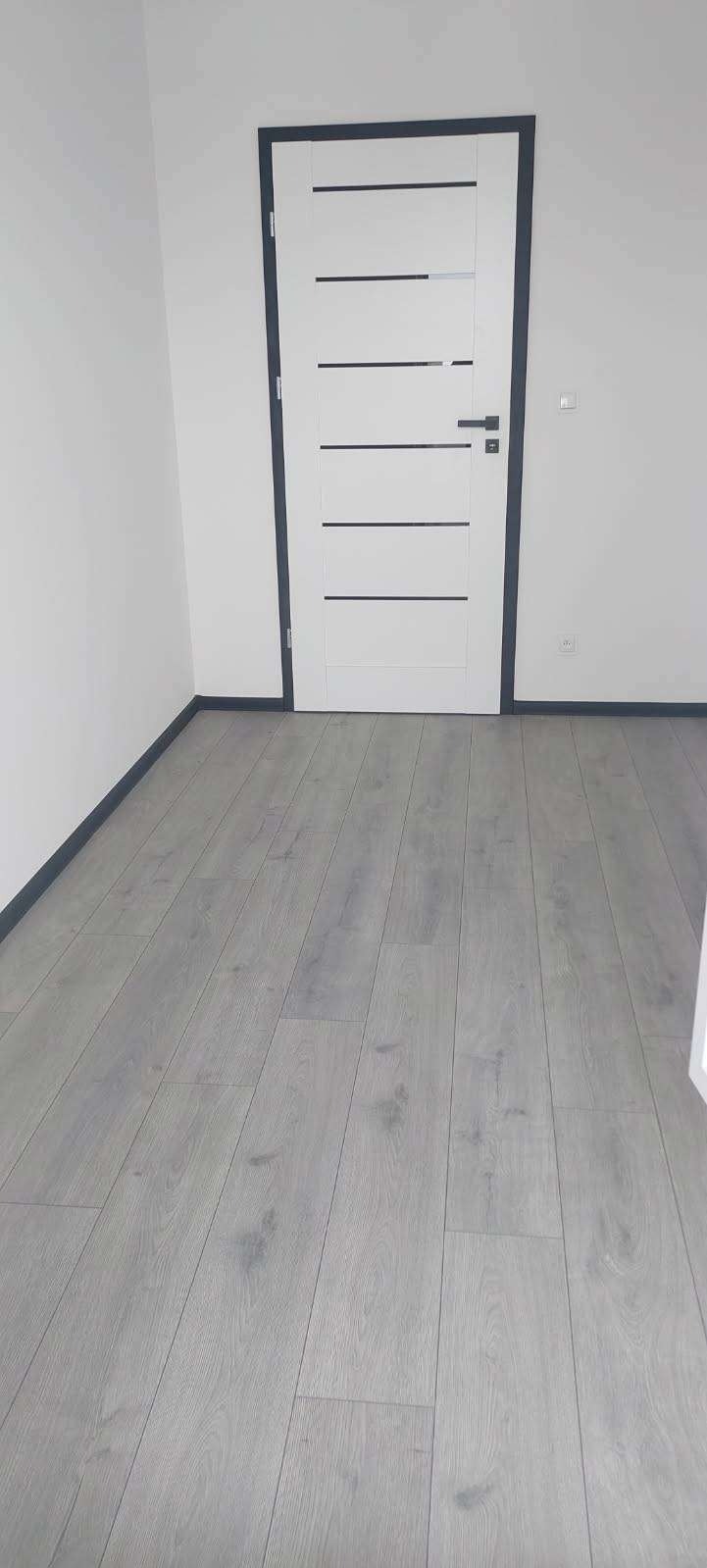 Szare panele podłogowe ułożone w jasnym pokoju z białą ścianą i białą, nowoczesną, wewnętrzną stolarką drzwiową z czarnymi akcentami.