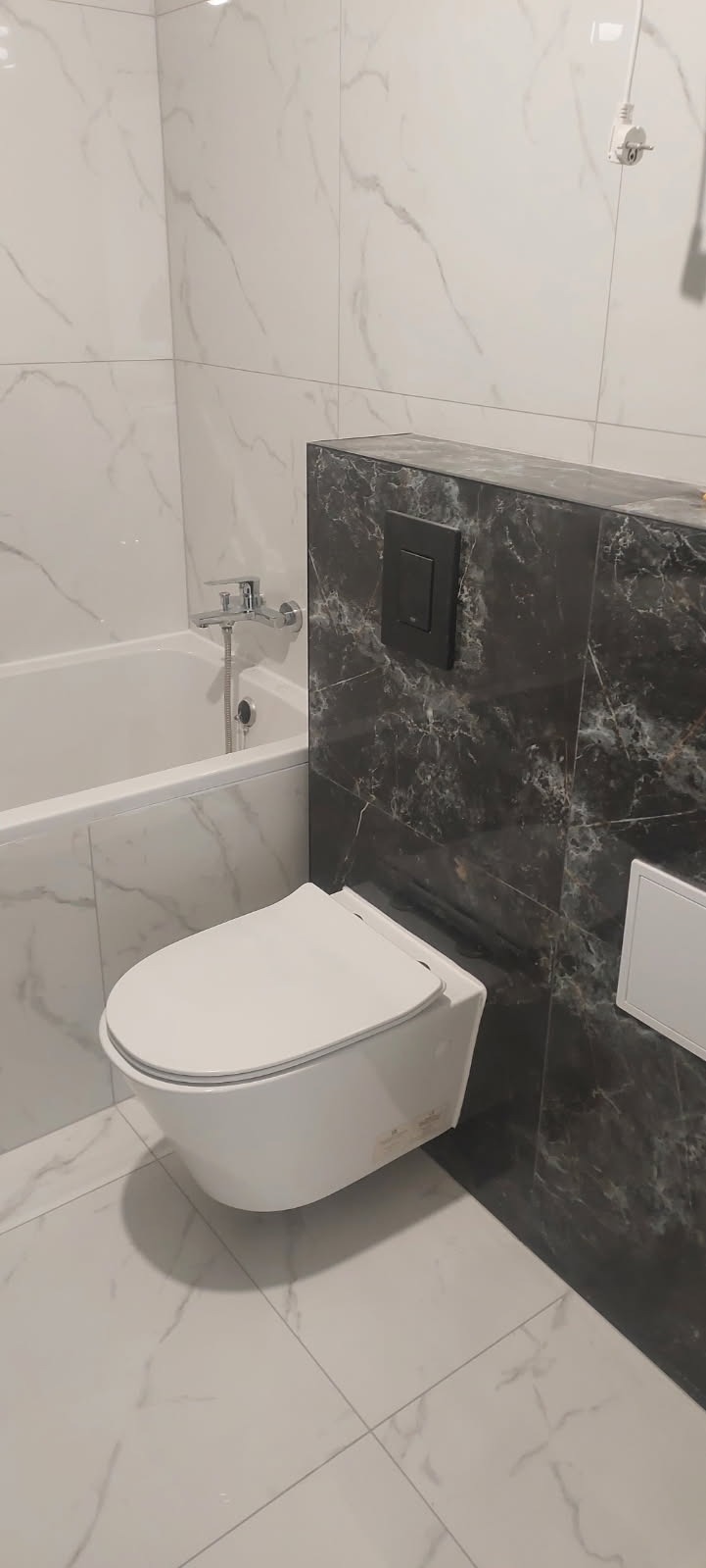 Nowoczesna, podwieszana toaleta z czarnym marmurem na zabudowie stelaża, białe płytki imitujące marmur na ścianach i podłodze, bateria wannowa chromowana.