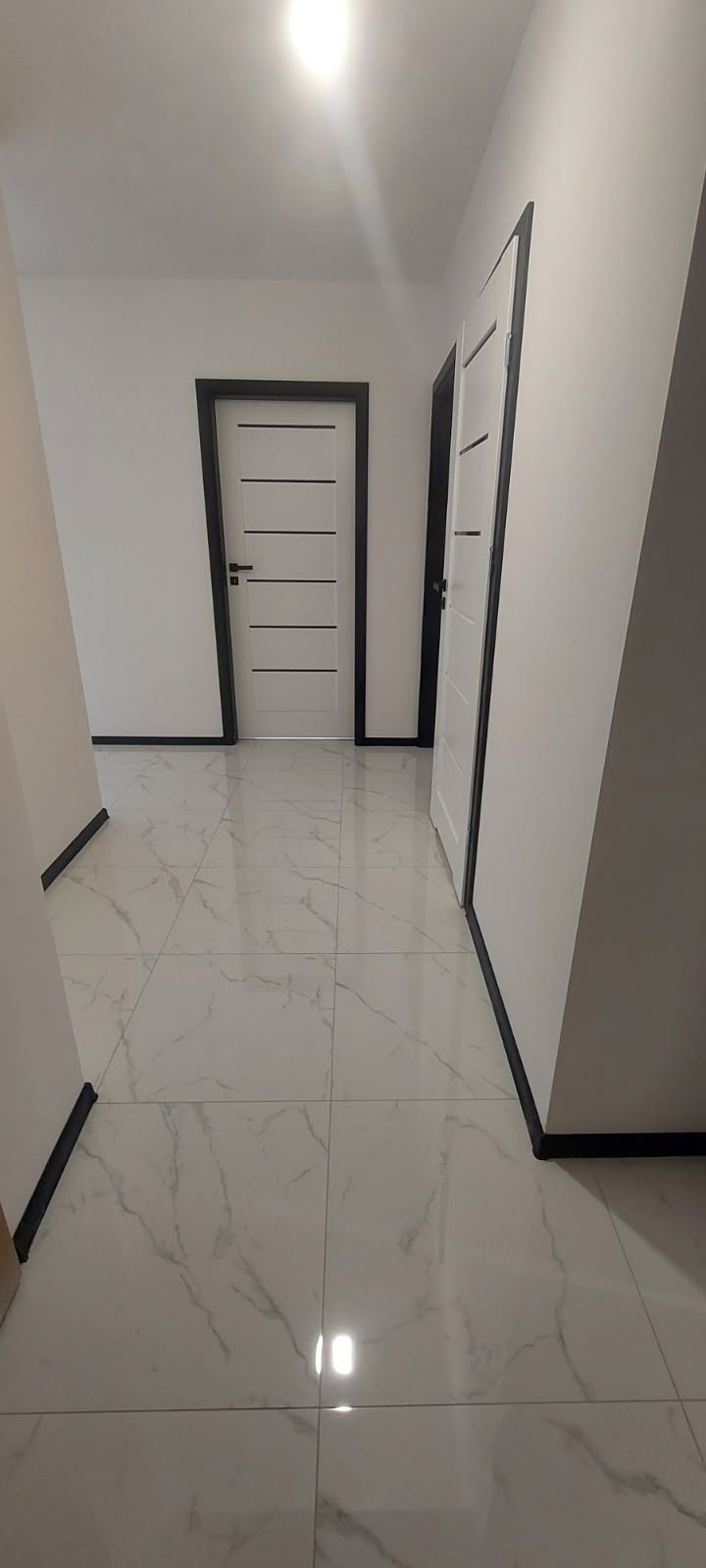 Korytarz z białymi ścianami, białymi płytkami podłogowymi imitującymi marmur i białymi drzwiami z czarnymi ościeżnicami oraz listwami przypodłogowymi.