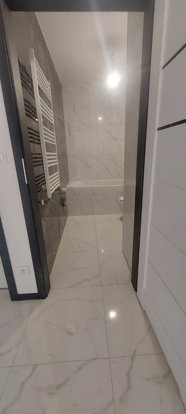 Nowoczesna łazienka z marmurową podłogą i ścianami, widoczny grzejnik drabinkowy, biała wanna i toaleta, ujęcie z perspektywy wejścia przez drzwi.