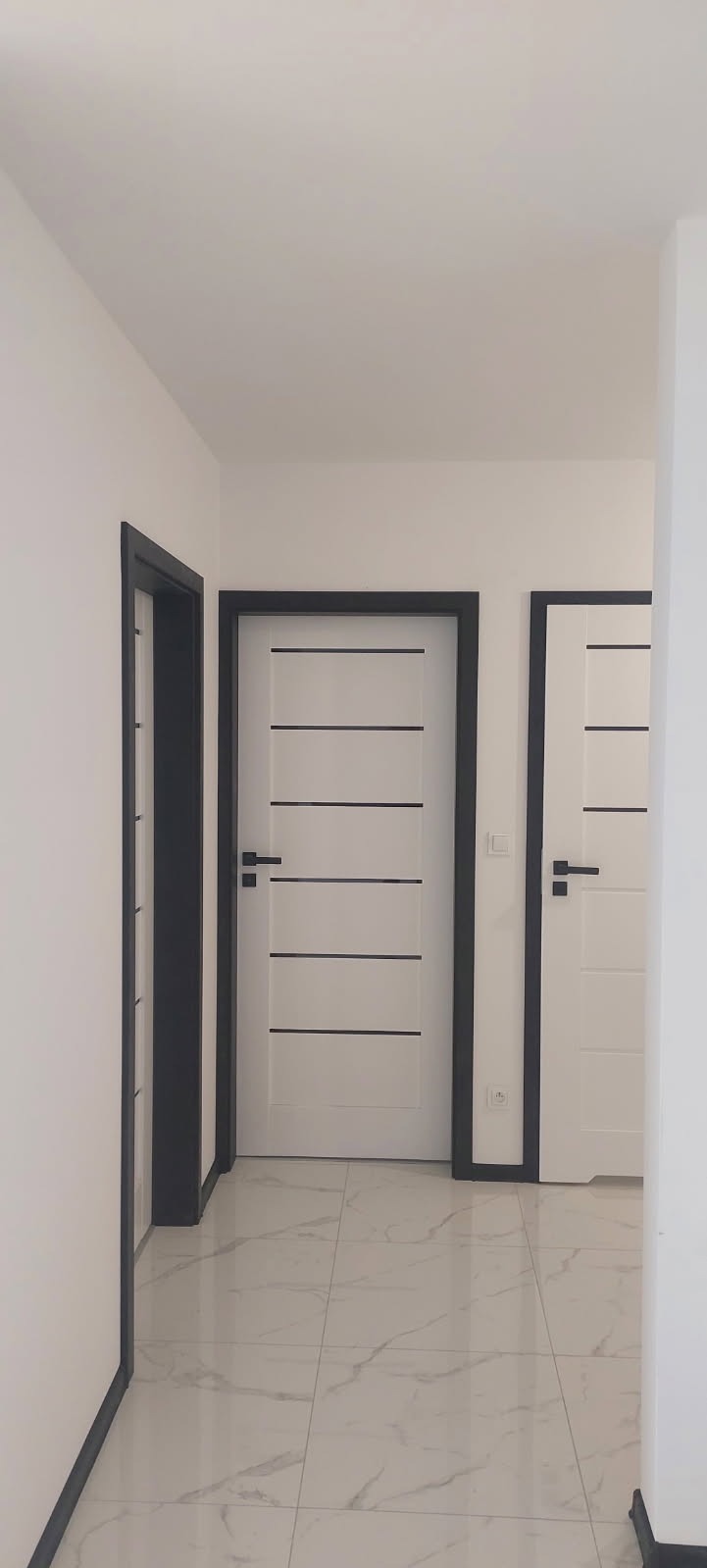 Korytarz z trzema białymi drzwiami z czarnymi ramami i poziomymi paskami, wykończony płytkami imitującymi marmur.
