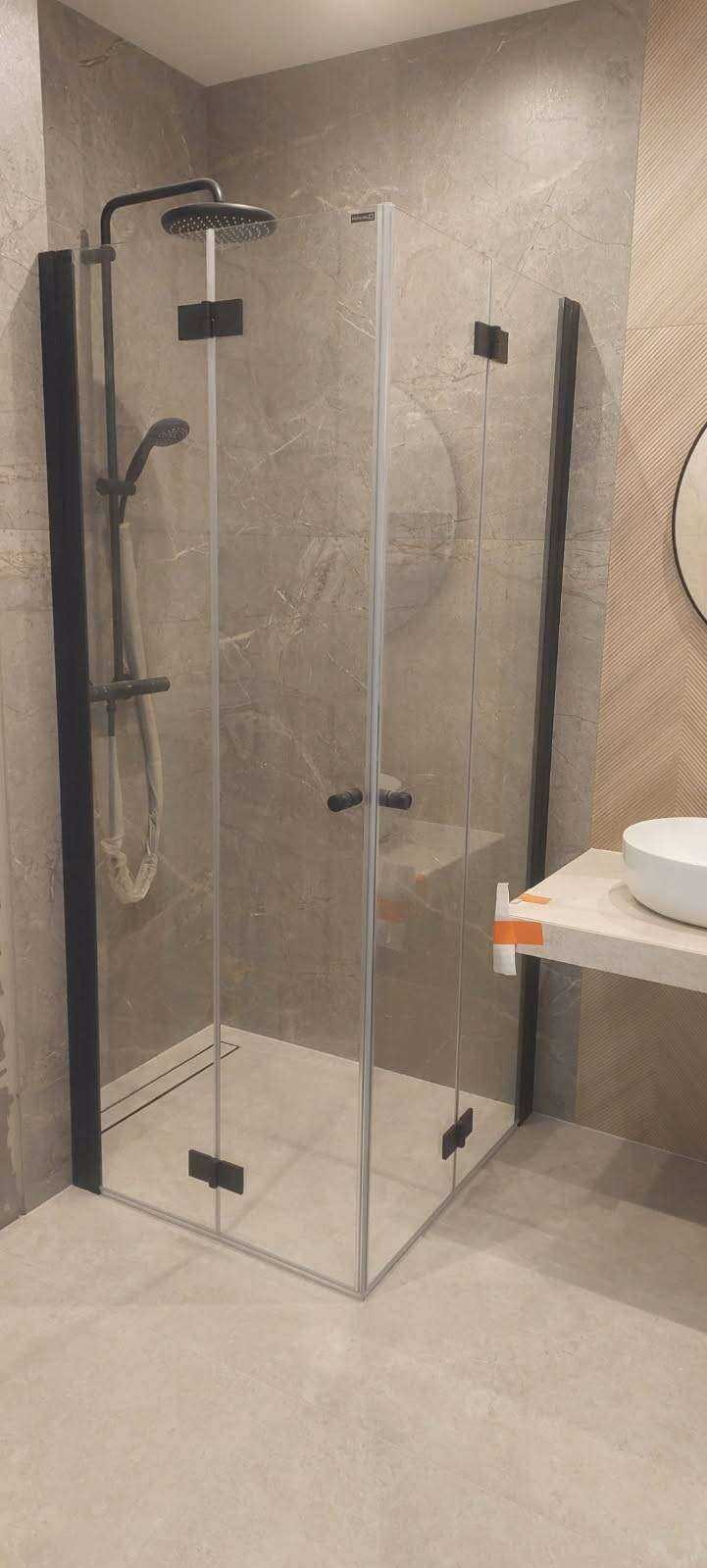 Nowoczesna kabina prysznicowa z czarnymi profilami i deszczownicą, zamontowana w łazience wykończonej płytkami imitującymi marmur. Na blacie obok kabiny widoczna biała umywalka.