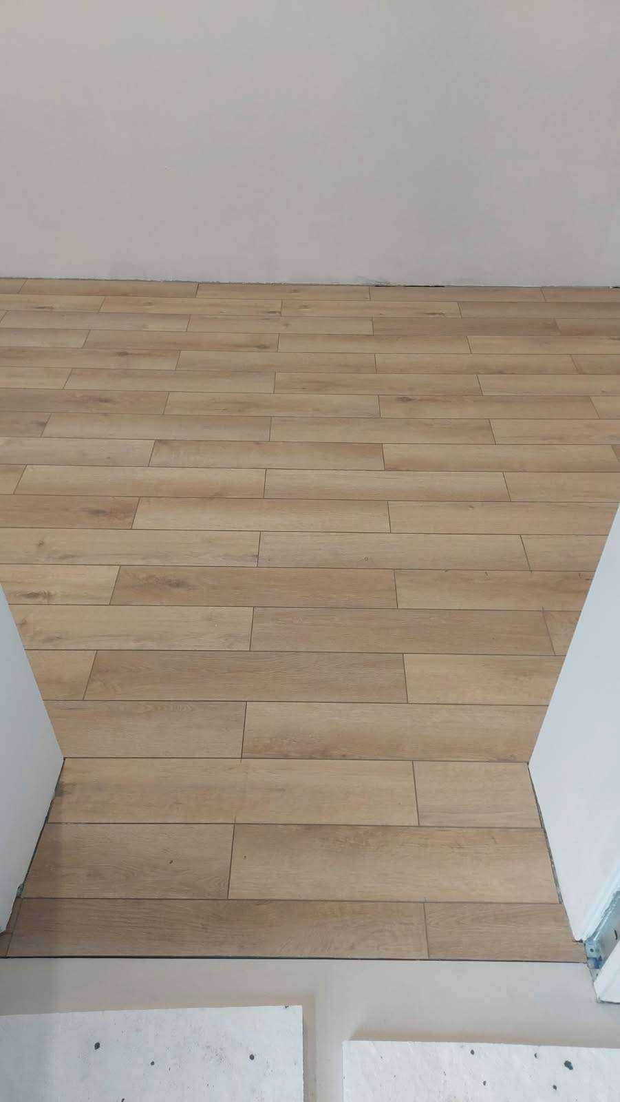 Ułożona podłoga z paneli imitujących drewno, widok z góry na fragment pomieszczenia z białymi ścianami i surowym betonem przy progu.