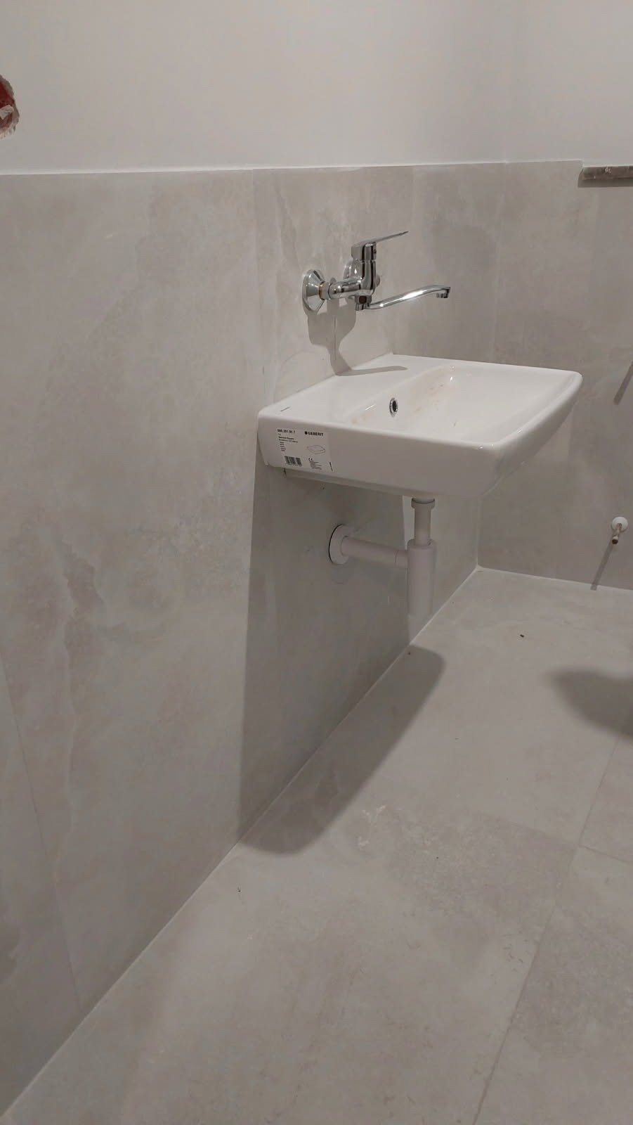Biała umywalka ceramiczna zamontowana na ścianie wyłożonej płytkami imitującymi marmur, widoczna bateria chromowana i syfon, na ścianie ślady po niedokończonych pracach instalacyjnych.