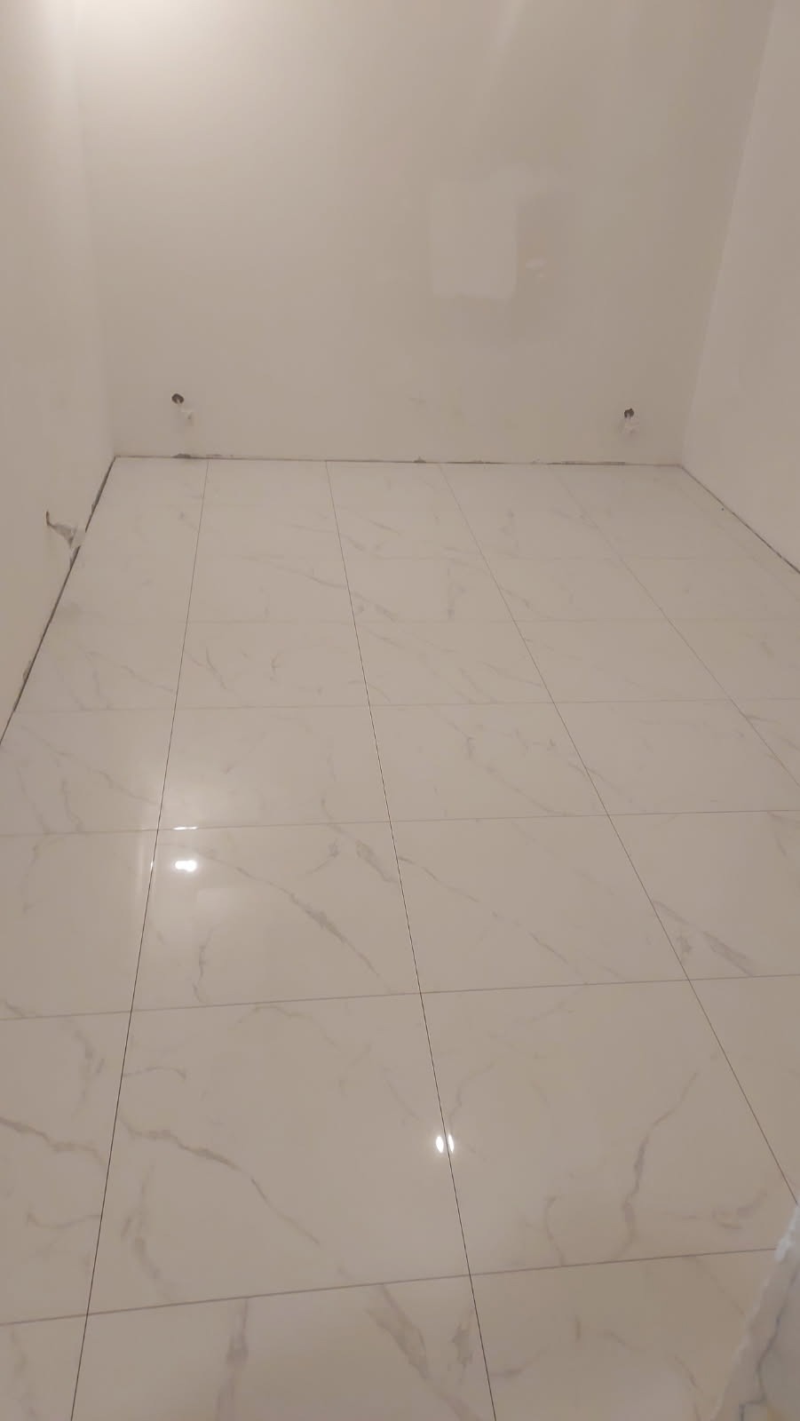 Wykończony pokój z białymi, marmurkowymi płytkami podłogowymi, z widocznymi przyłączami do armatury na ścianie.