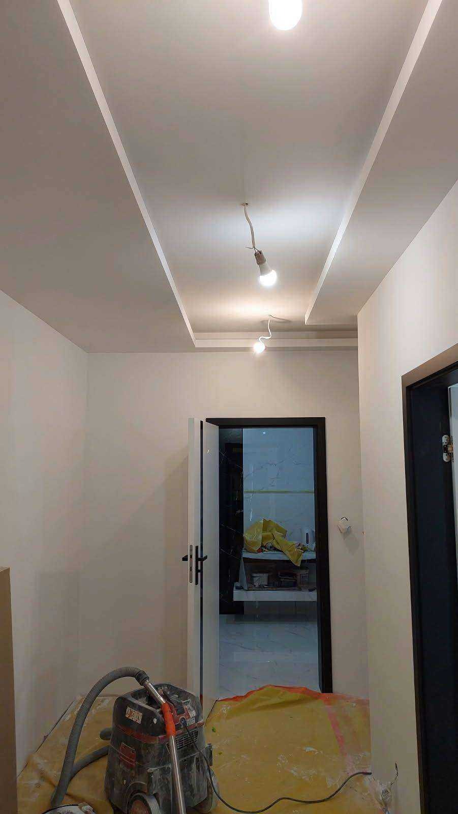 Korytarz w trakcie prac wykończeniowych: świeżo pomalowane ściany, sufit z dekoracyjnymi listwami i zamontowanym oświetleniem, zabezpieczona podłoga oraz odkurzacz przemysłowy.