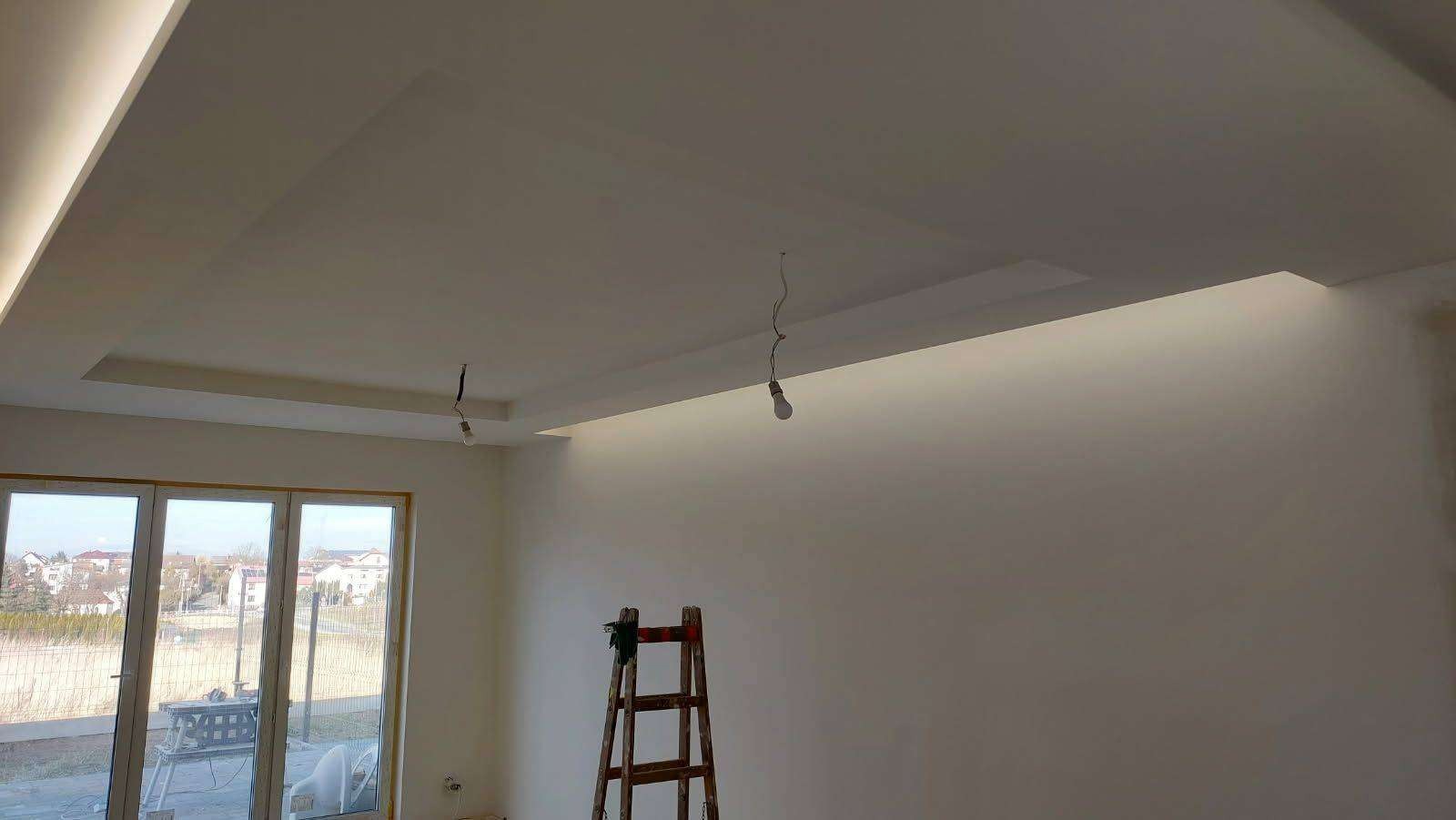 Wnętrze pokoju z pomalowanymi na biało ścianami i sufitem, z widocznymi elementami oświetlenia w suficie podwieszanym i wiszącymi żarówkami na kablach, drewniana drabina oparta o ścianę.
