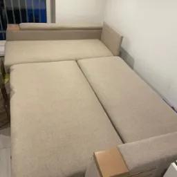 Beżowa sofa modułowa z drewnianymi akcentami, rozłożona, widoczna w pomieszczeniu z oknem i białą ścianą.