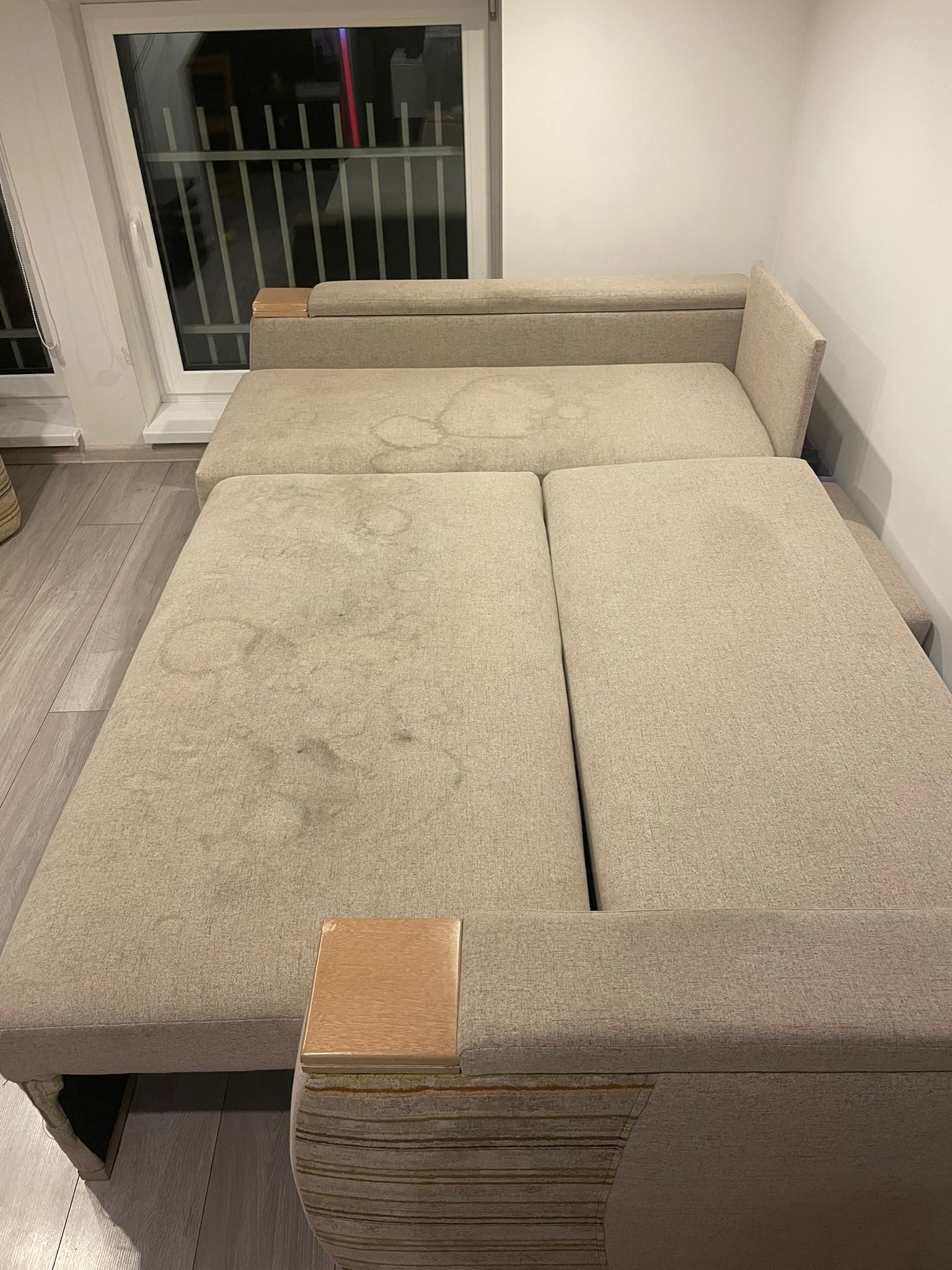 Rozłożona sofa w odcieniach beżu z widocznymi, zaschniętymi plamami na siedzisku, sfotografowana w słabym oświetleniu wnętrza mieszkalnego.