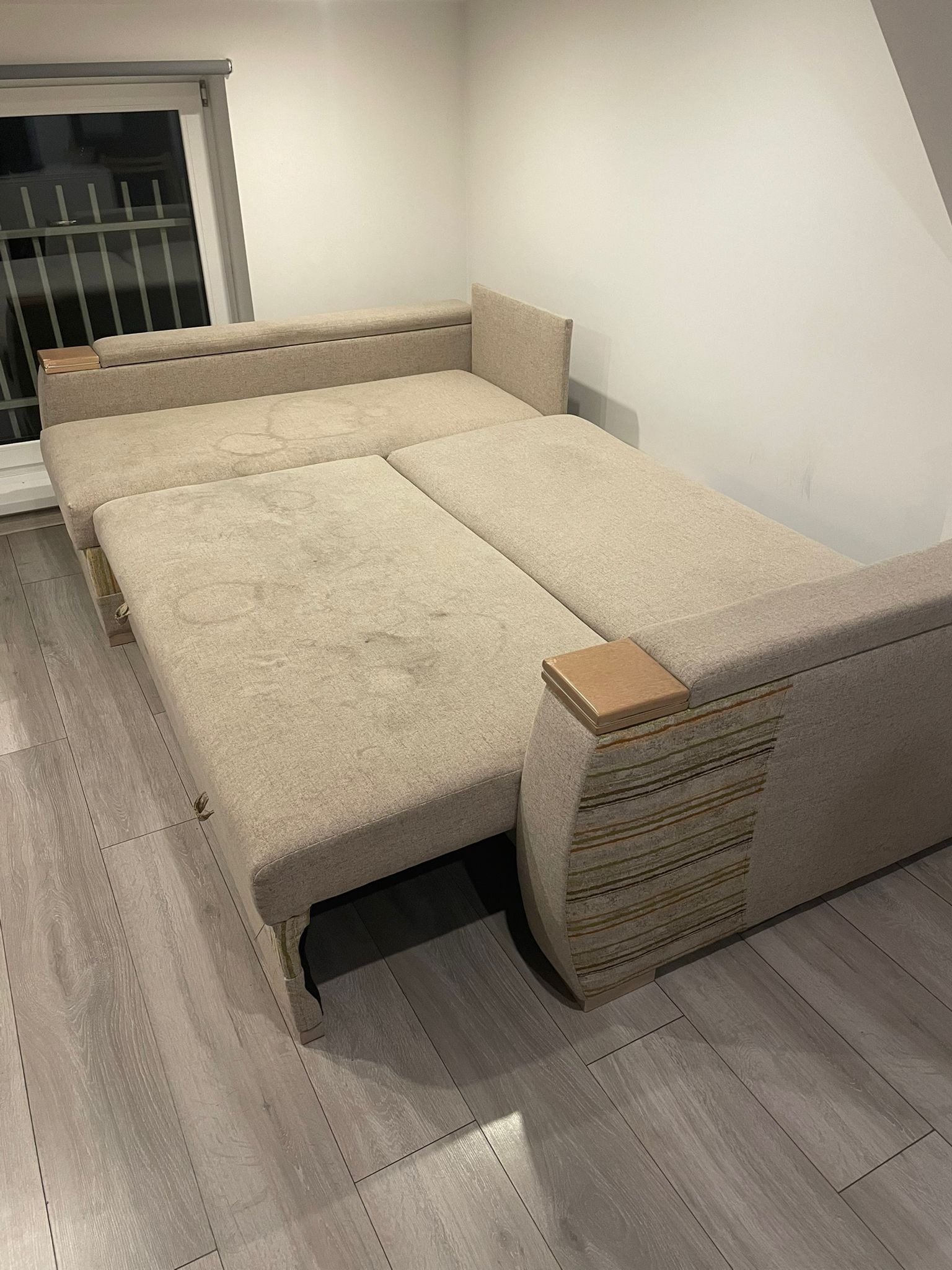 Rozłożona, beżowa sofa z widocznymi plamami na tapicerce, ustawiona na panelach podłogowych, z oknem w tle.