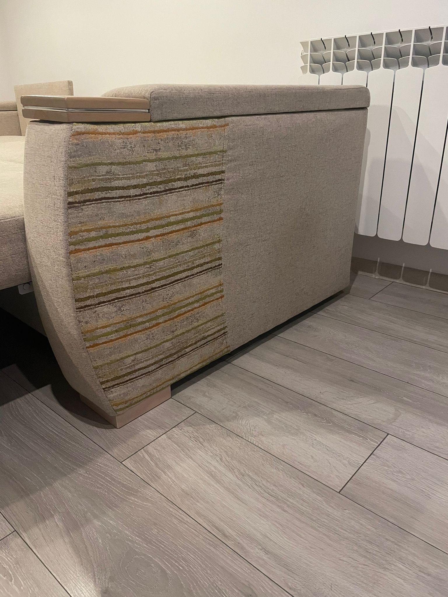 Fragment beżowej sofy z wzorzystym bokiem, widoczny na tle białego grzejnika i paneli podłogowych z imitacją drewna.