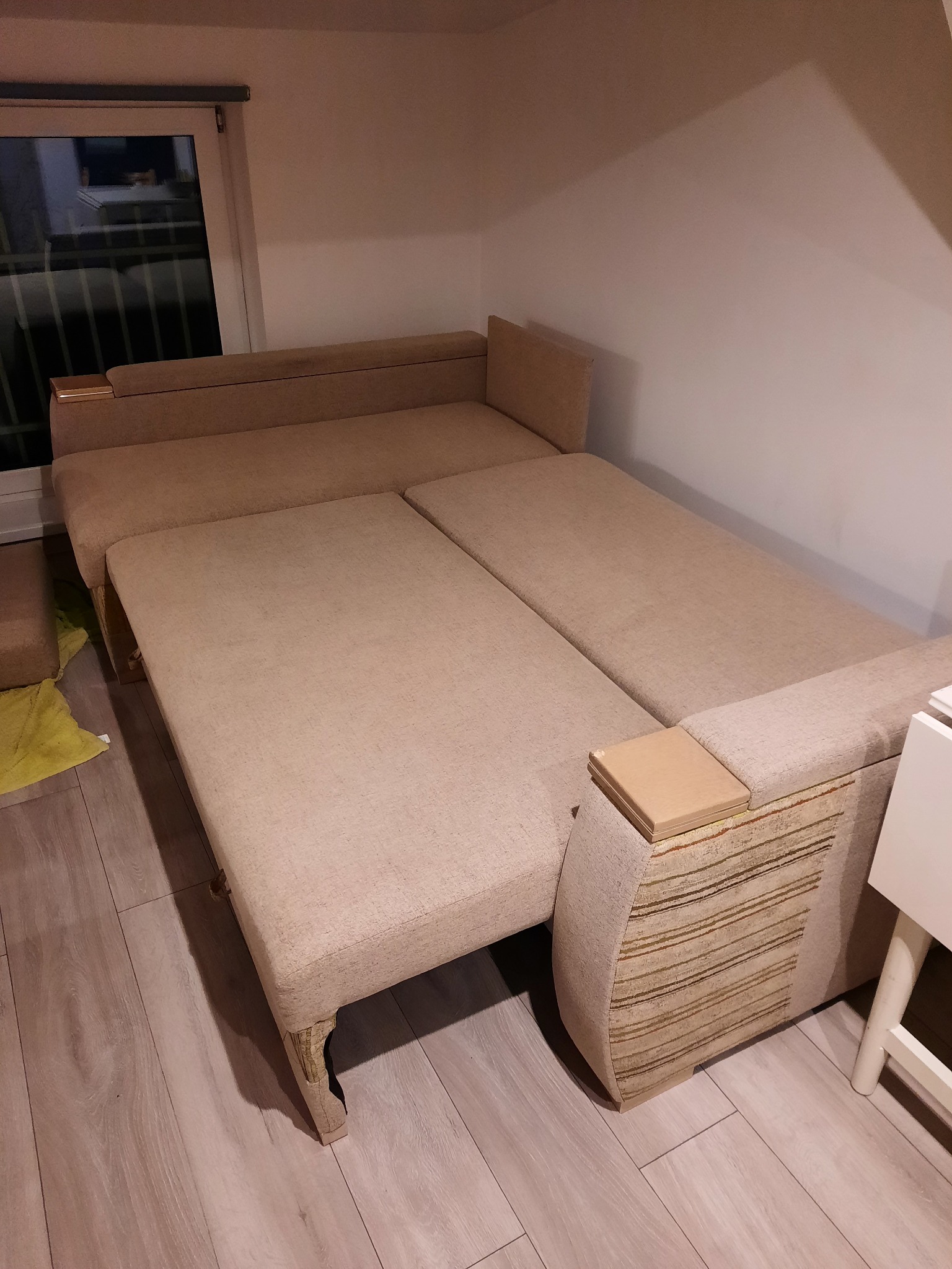 Rozłożona, beżowa sofa z widocznymi elementami dekoracyjnymi na bokach, stojąca na panelach podłogowych, obok biały stolik.